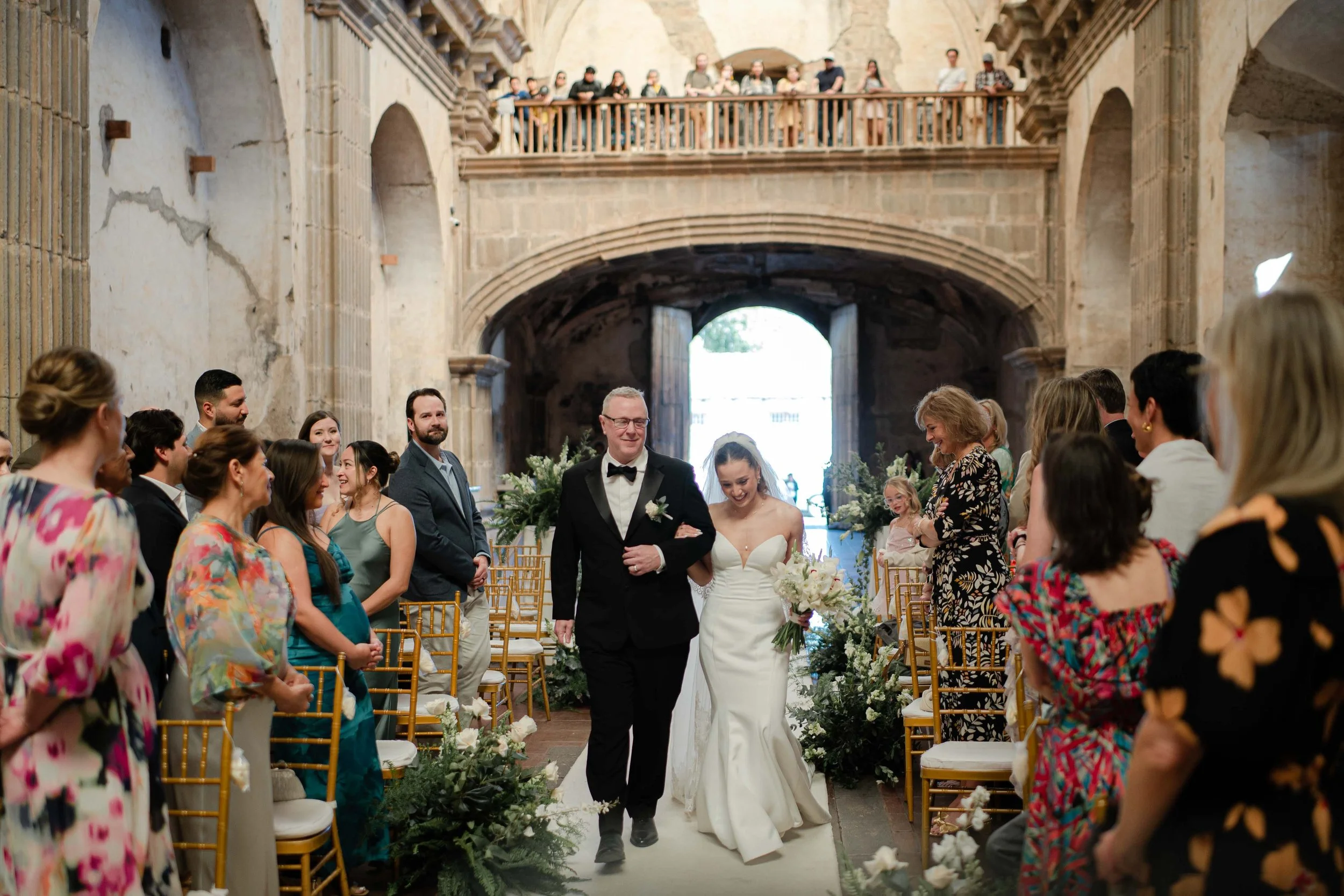 Pareja de recién casados caminando por pasillo en iglesia, rodeados de invitados vestidos con ropa formal, durante ceremonia de boda.