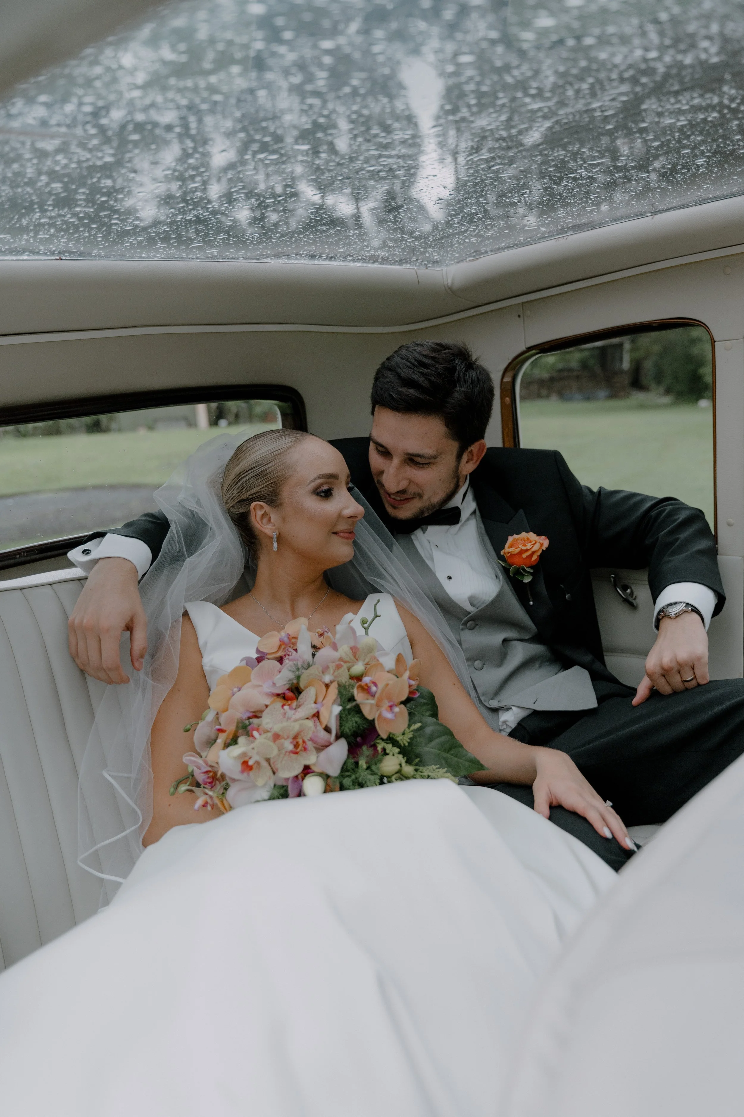 Una pareja de recién casados en el interior de un vehÃculo clásico, ella con vestido de novia y ramo de flores, él con traje y corbata, compartiendo un momento romántico