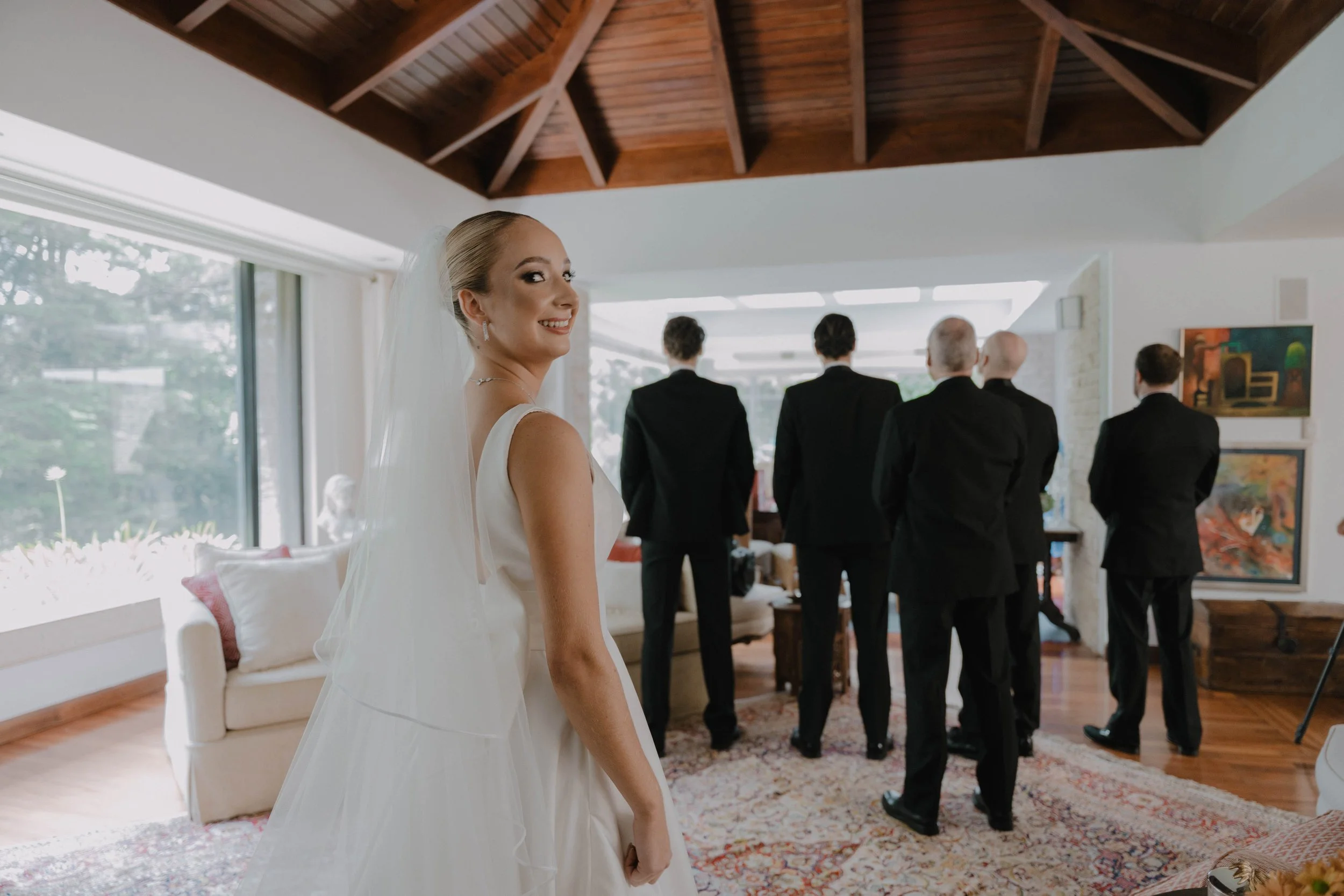 Una novia sonriendo en su vestido blanco de boda, mirando hacia la cámara, mientras en el fondo varios hombres de traje están de espaldas en una sala con ventanas grandes y arte en las paredes.