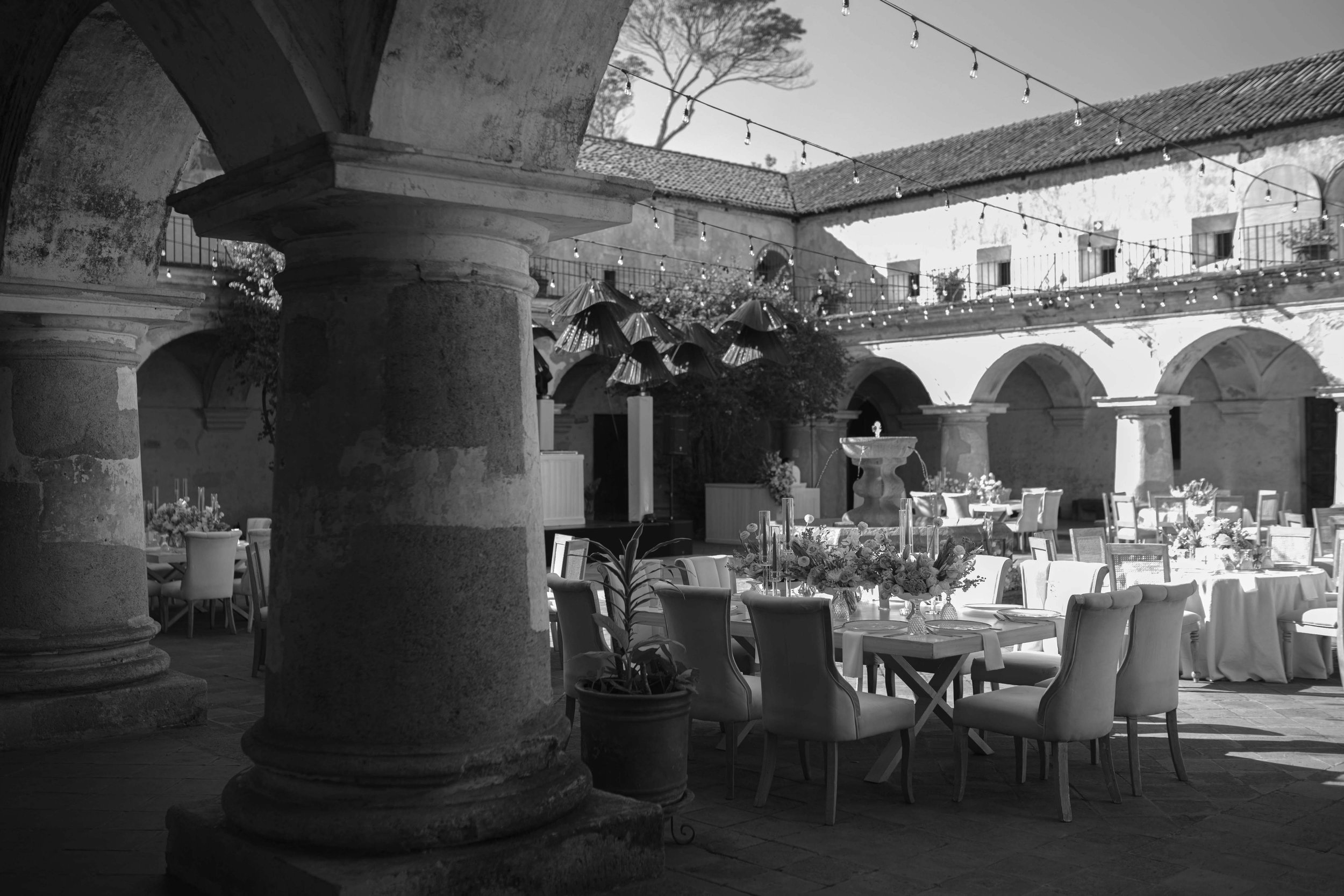 Cenador en un patio con mesas decoradas con flores para un evento, con columnas, paredes de piedra, y luces colgantes