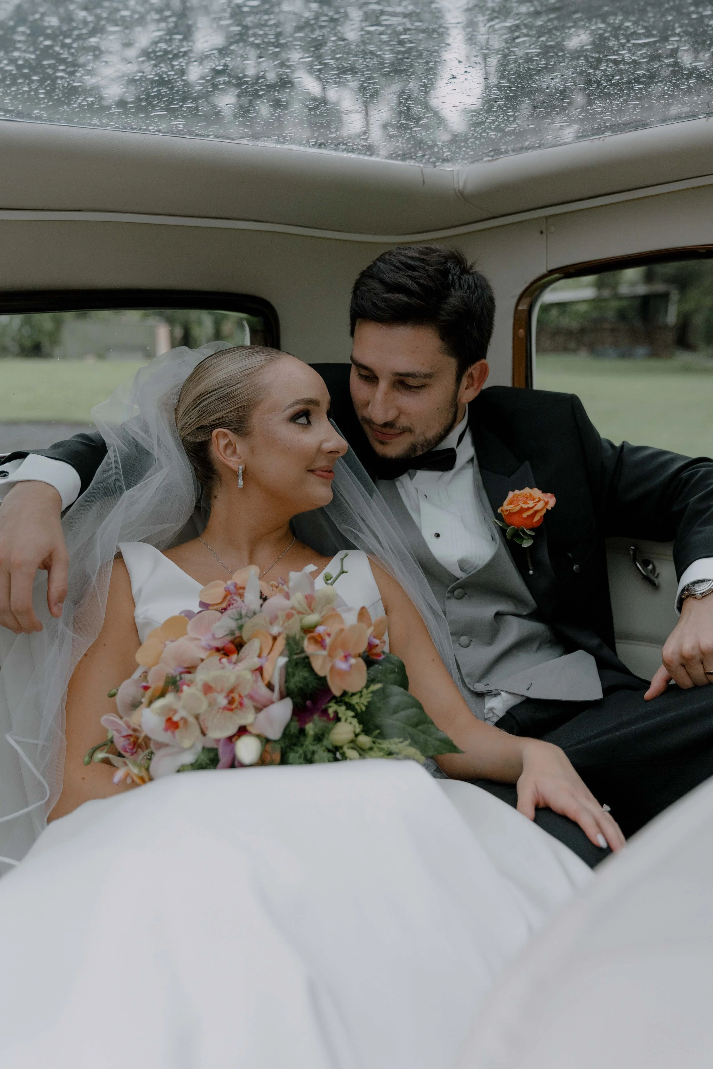 Pareja de recién casados en un coche, ella con vestido de boda y ramo de flores, él vestido con esmoquin y flor en el ojal, compartiendo un momento romántico