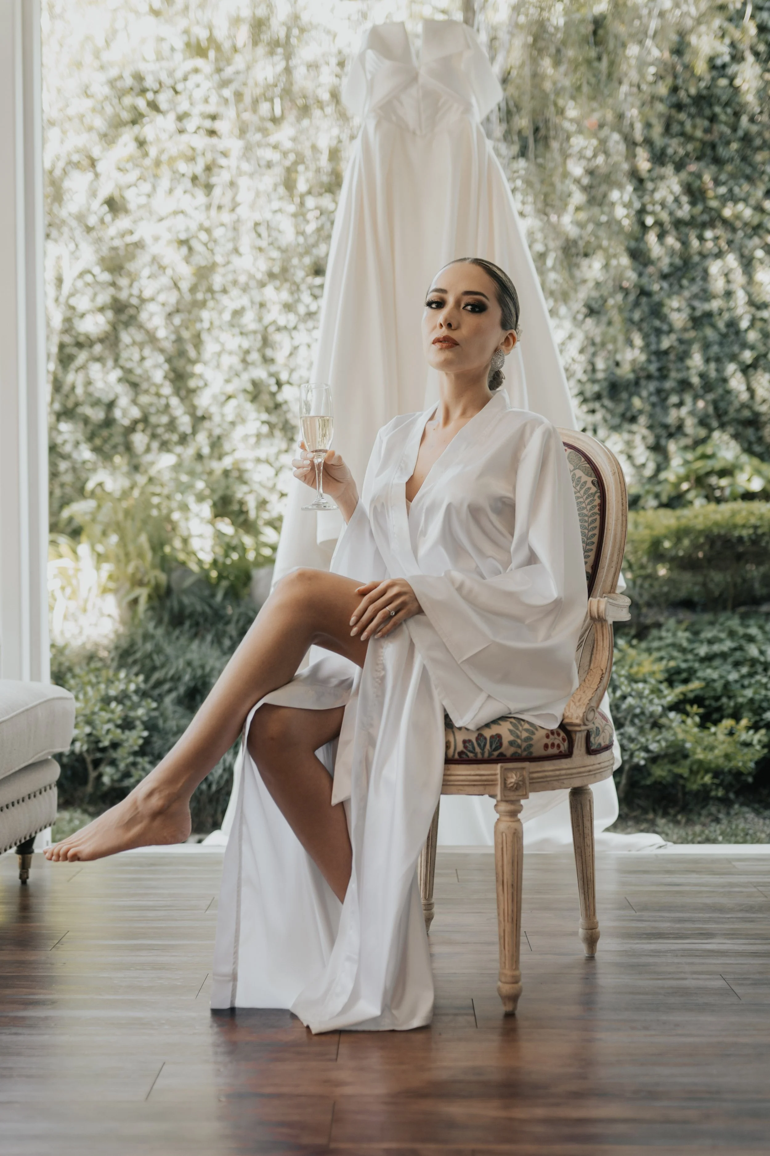 Mujer en bata satinada blanca sentada en una silla, con copa de champagne en mano, detrás una vestido de novia colgado en una ventana con paisaje natural exterior.