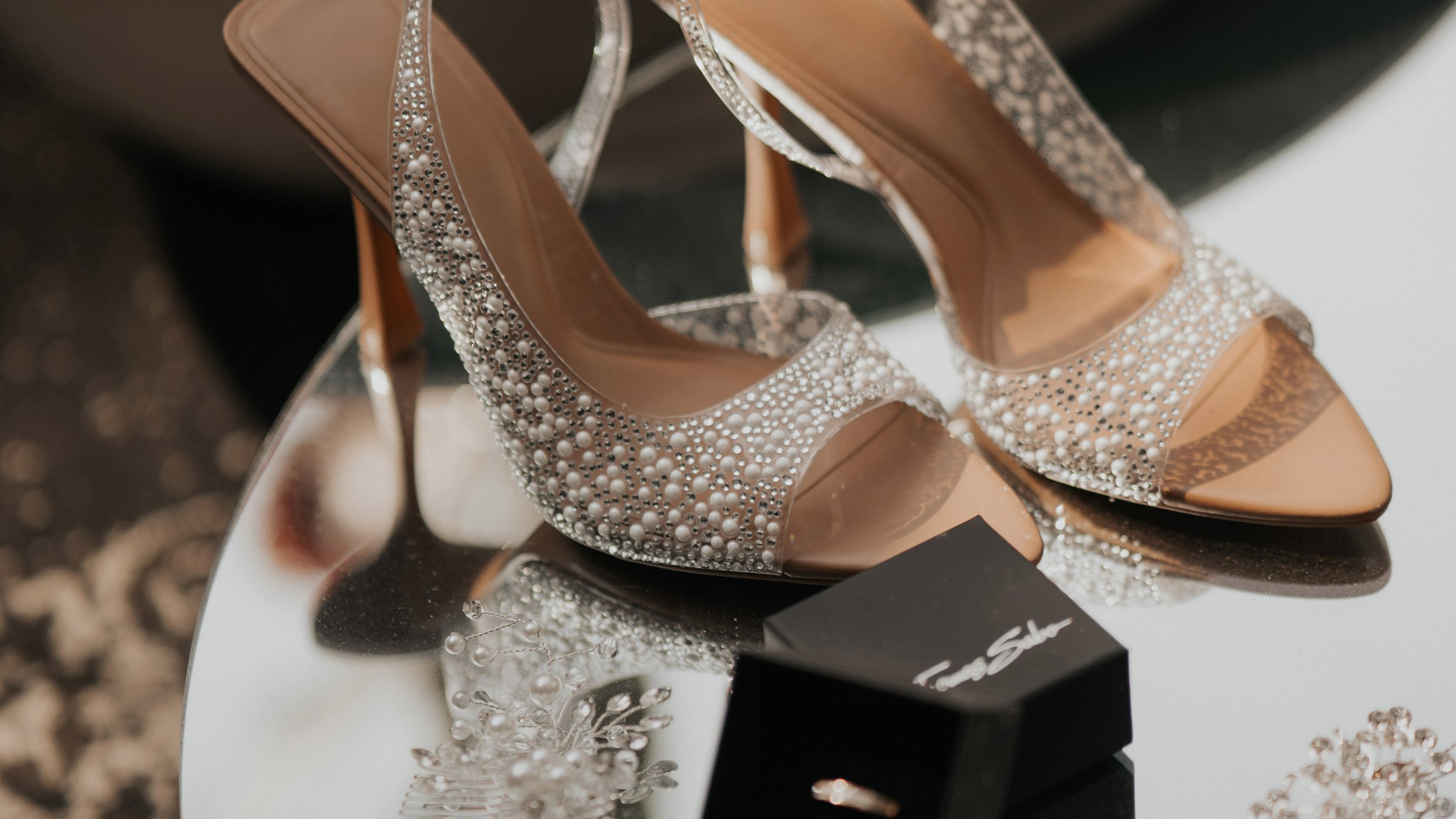 Zapatos de tacón con detalles de perlas y cristales en una mesa, cerca de una caja negra y accesorios de joyería blancos y plateados.