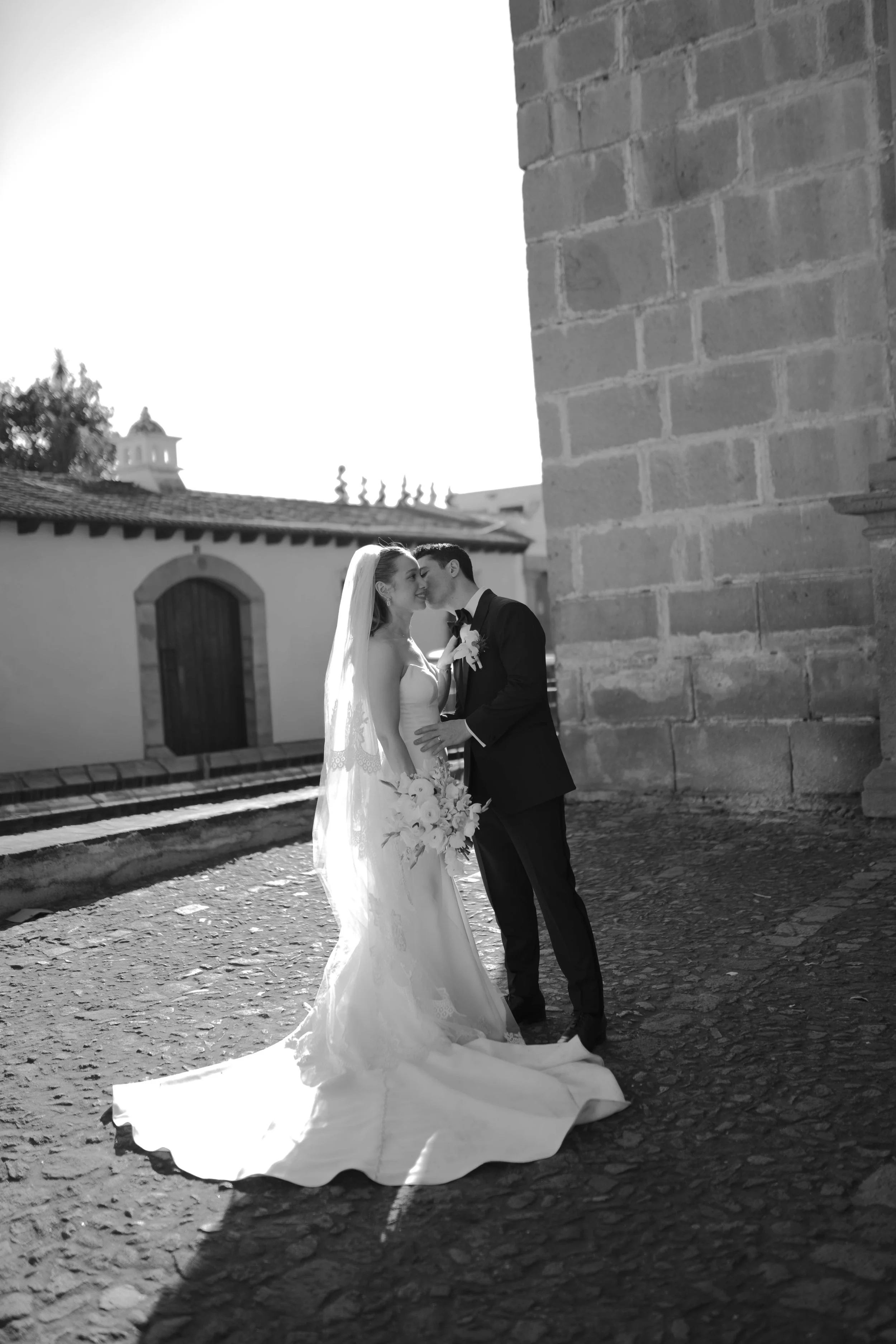 Pareja de recién casados, ella con vestido de novia y velo, él con traje formal, en un lugar al aire libre, abrazados y compartiendo un beso. La fotografía en blanco y negro.