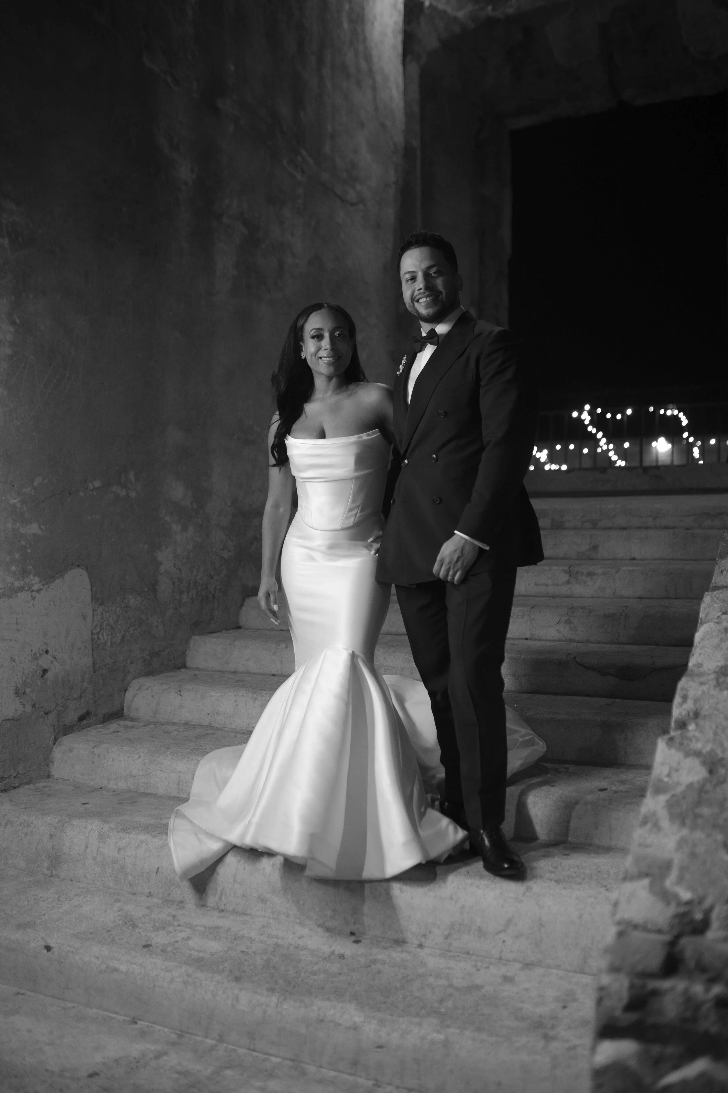 Una pareja vestida con ropa de boda, la mujer con vestido largo y el hombre con traje formal, posando en unas escaleras con una pared de fondo y luces decorativas en la distancia.