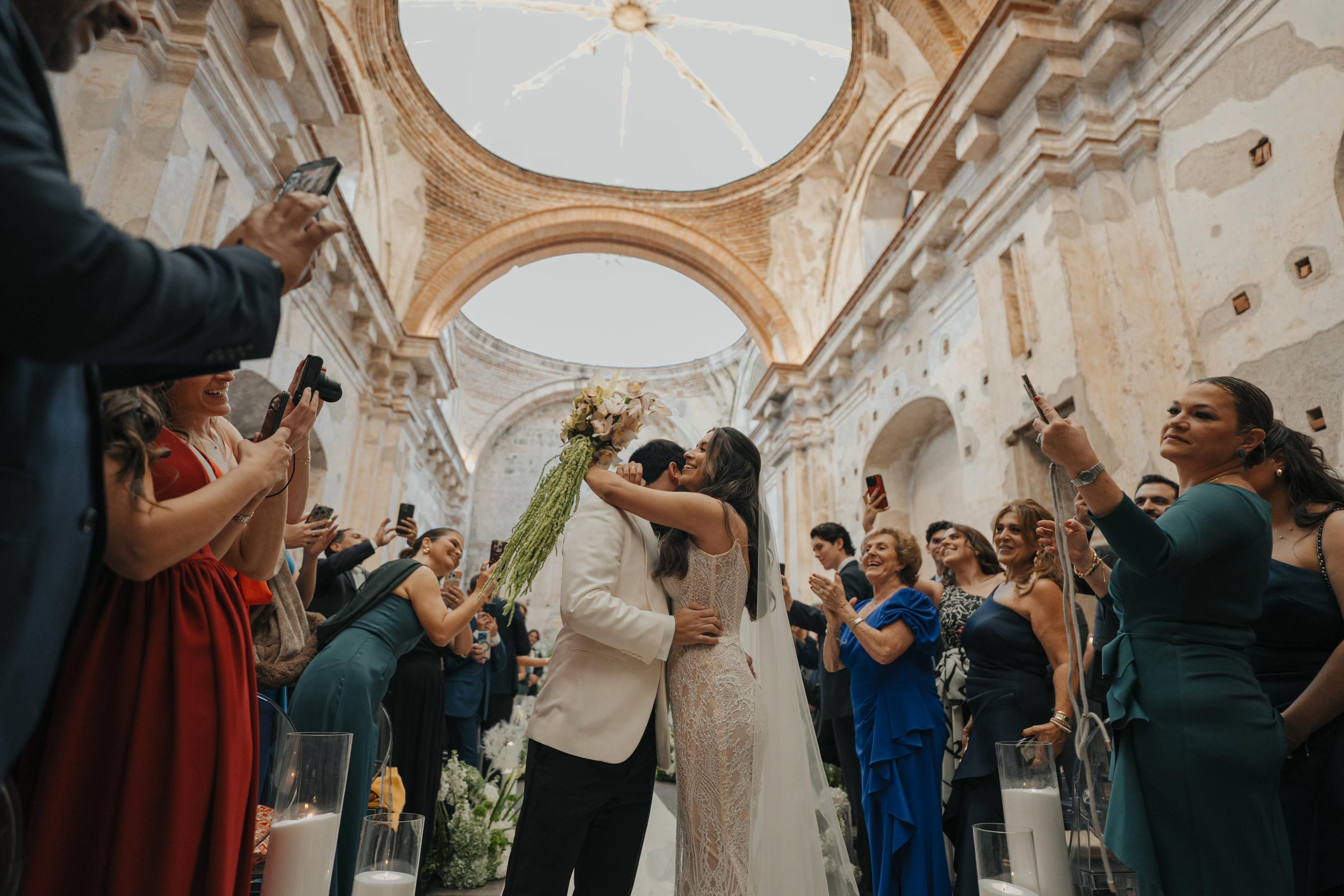 Pareja de recién casados abrazándose y besándose en una boda dentro de una iglesia antigua, rodeados de invitados que toman fotos