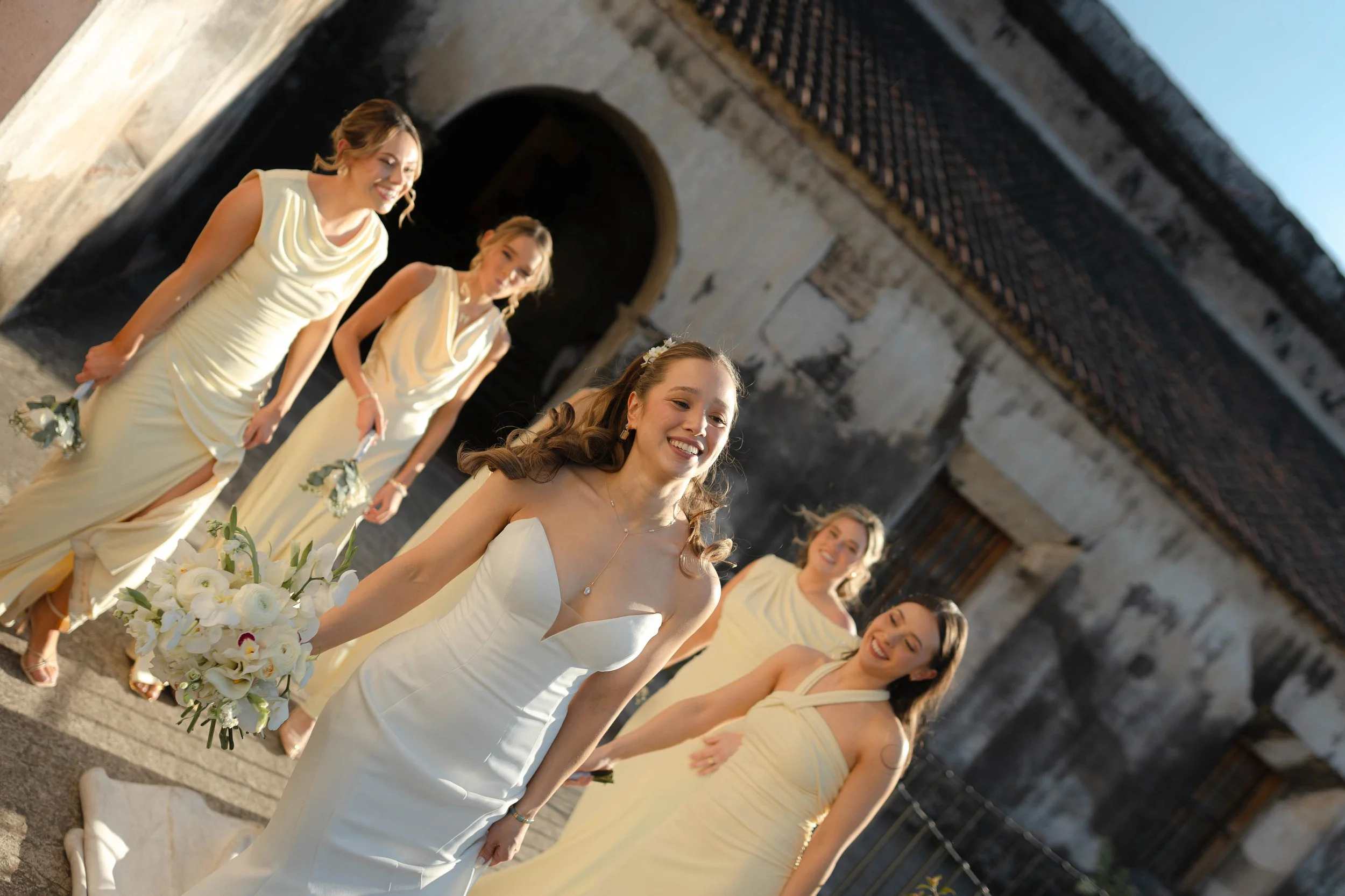 Grupo de mujeres en vestido de novia y damas de honor en una boda al aire libre frente a una estructura antigua de piedra.