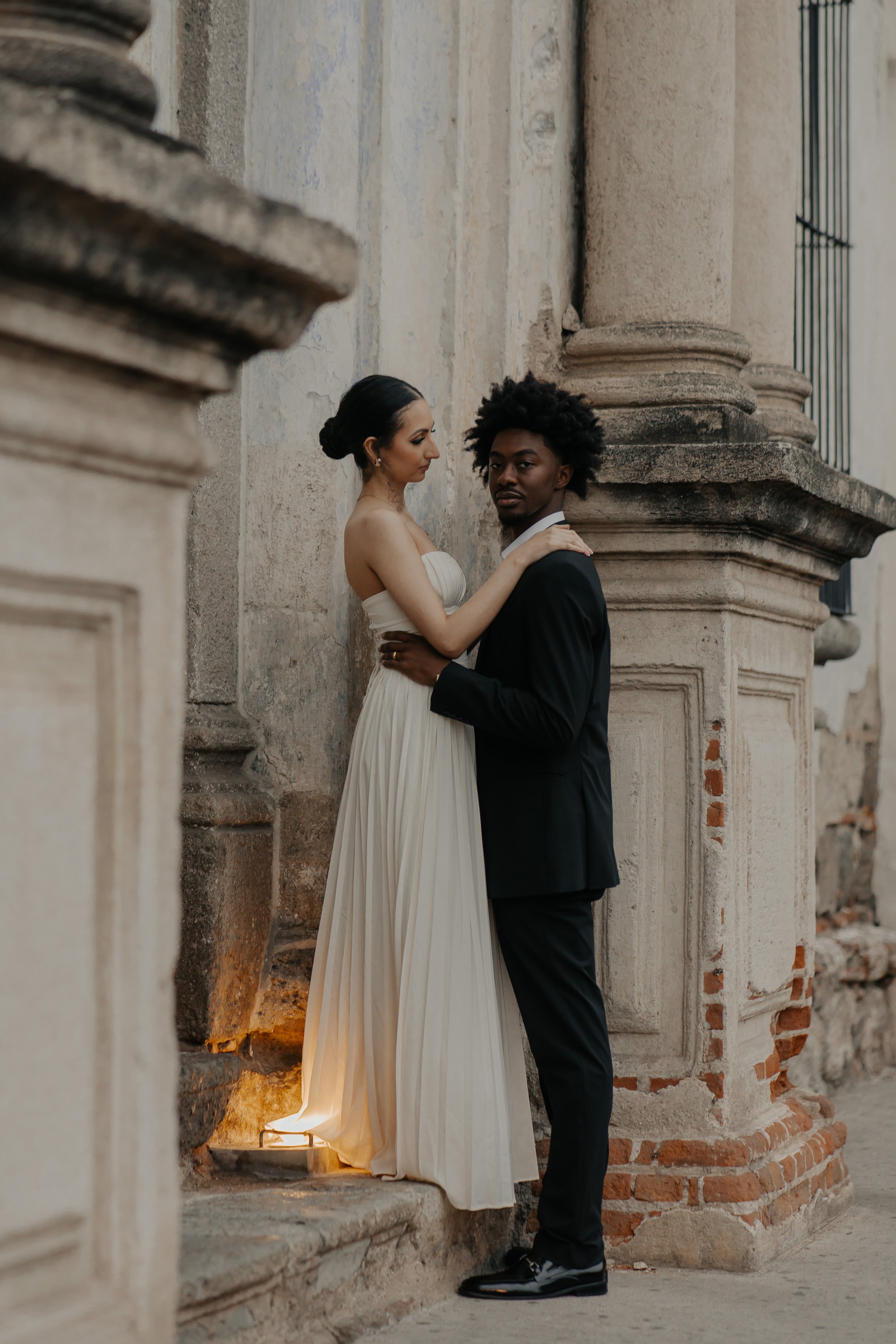 Una pareja vestida de boda, la mujer con vestido largo y el hombre con traje, frente a una pared vintage de piedra y ladrillo.