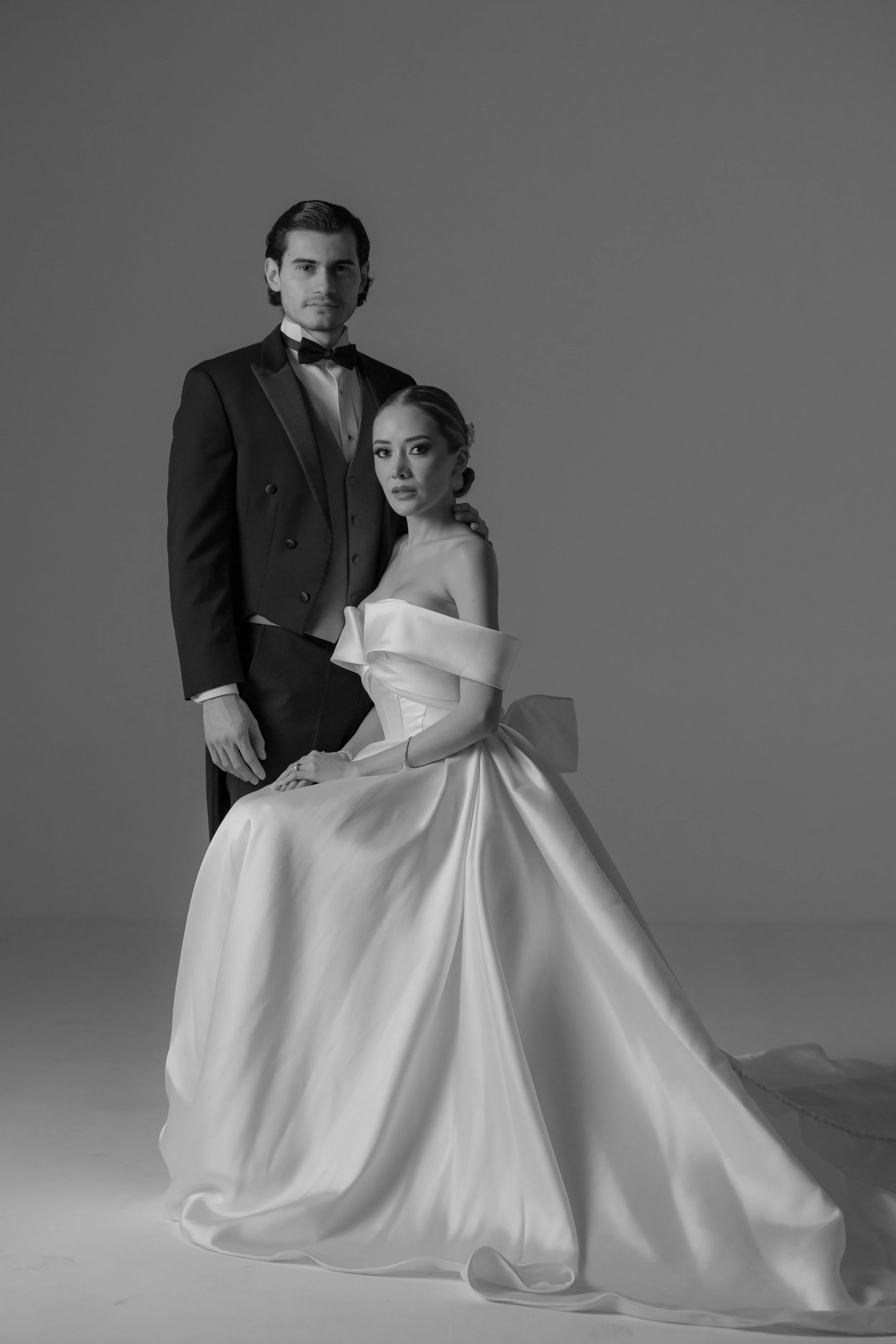 Una pareja vestida con ropa elegante en un estudio, una mujer en un vestido de novia y un hombre en un traje de tuxedo, en blanco y negro.