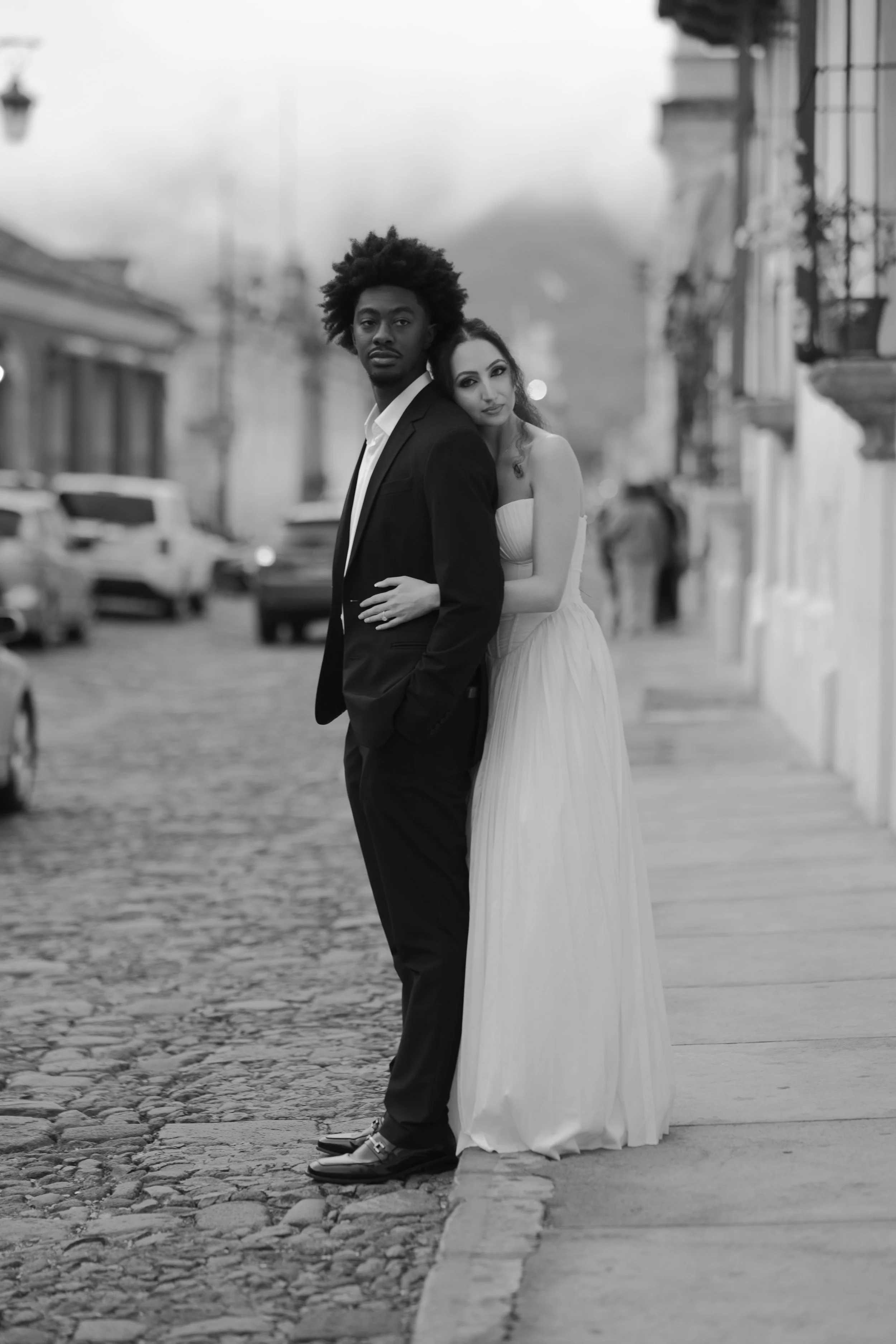 Pareja de esposos en un callejón empedrado, con mujer vestida de blanco y hombre de traje oscuro, en una escena en blanco y negro