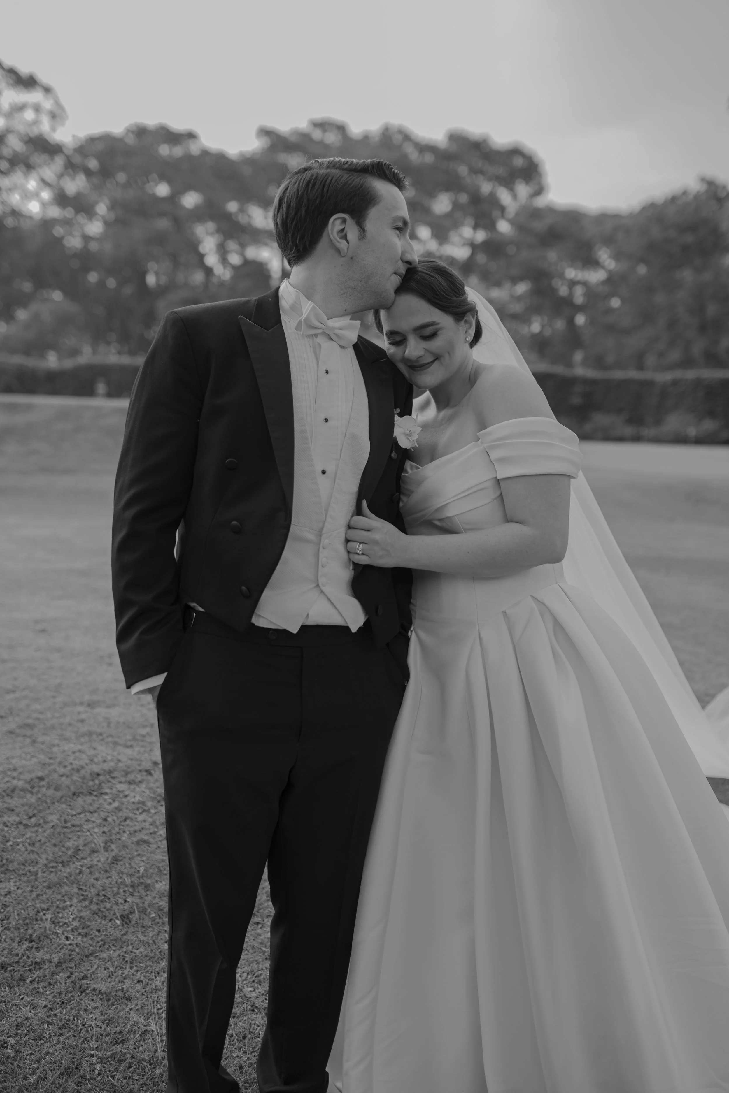 Pareja de novios en una boda, él en traje formal y ella en vestido de novia, en un campo con árboles de fondo, en blanco y negro.