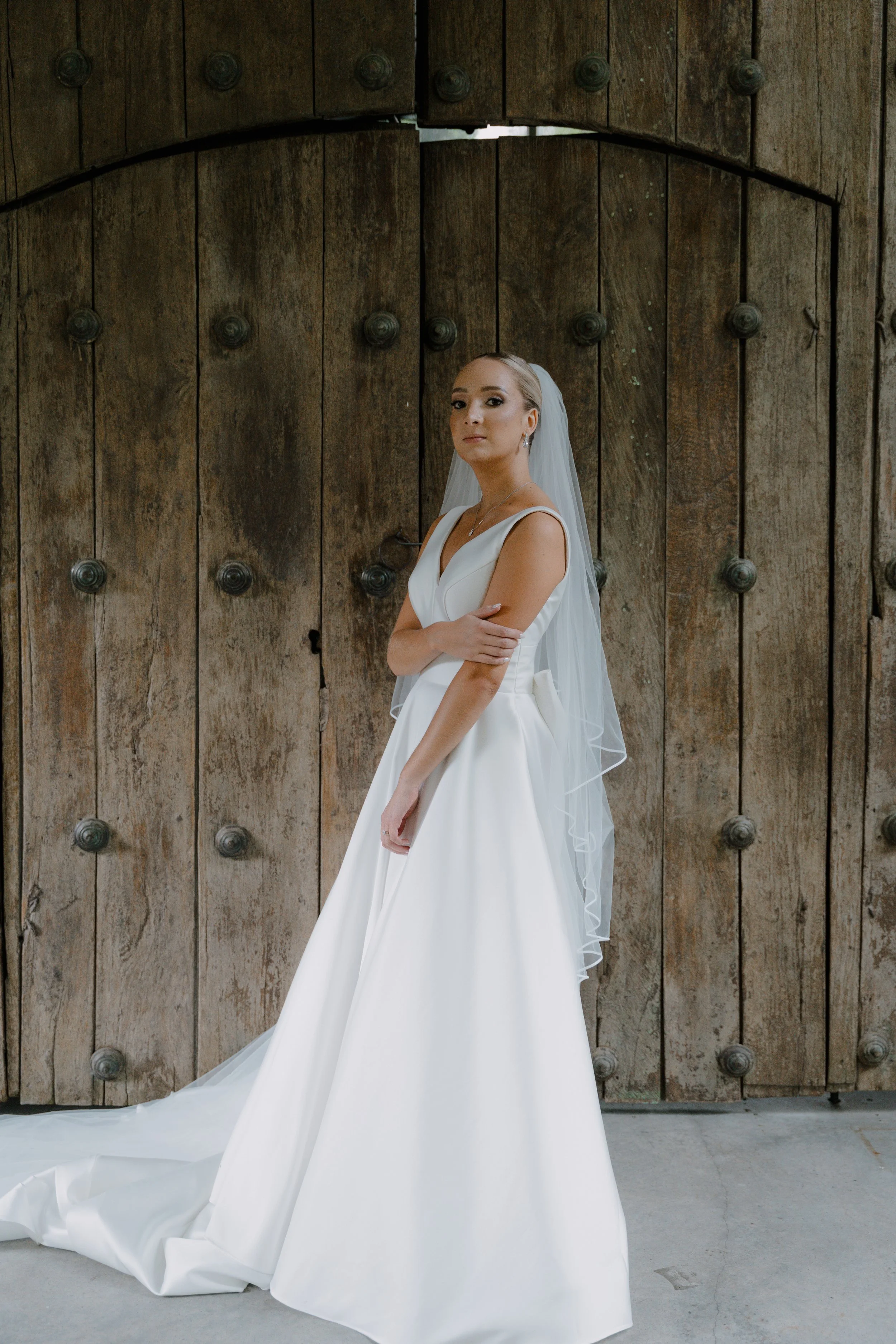 Mujer en vestido de novia blanco y velo, posando frente a una puerta de madera grande y antigua.