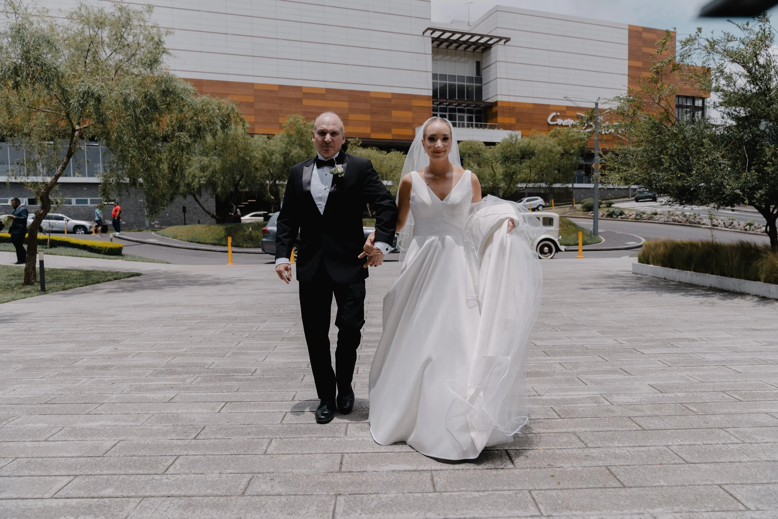 Pareja de novios caminando en un espacio abierto, la novia con vestido de novia y el novio con saco y pajarita, en un dÃa soleado con árboles y edificios modernos en el fondo.