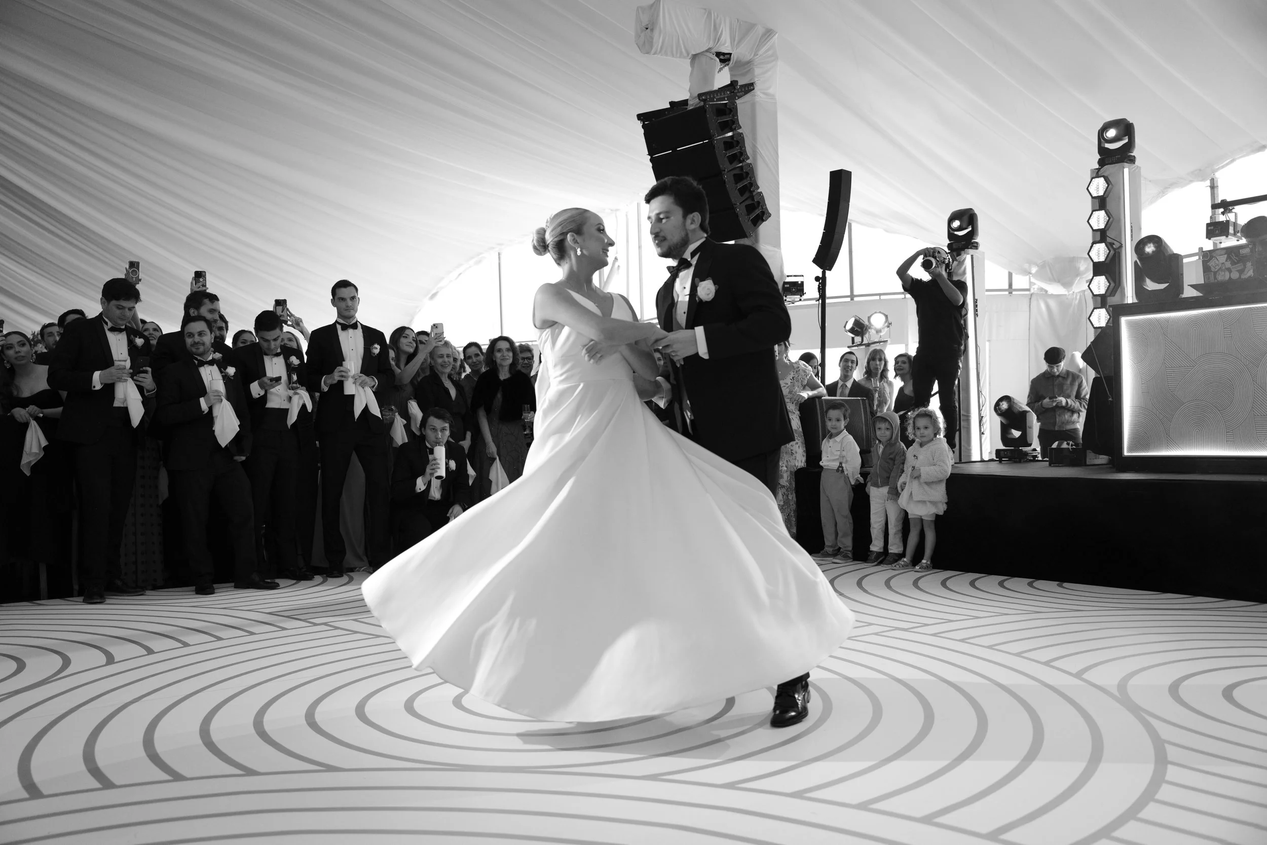 Pareja de novios bailando en su boda, rodeados de invitados en un lugar de ceremonia elegante.