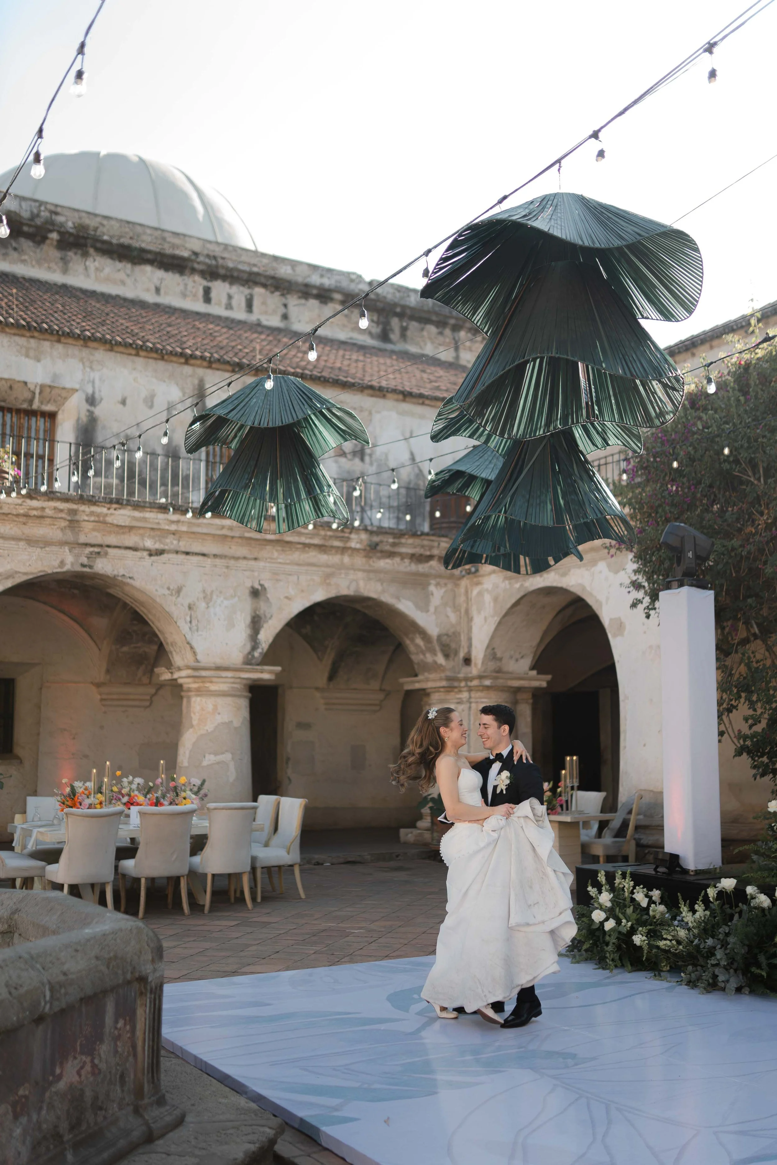 Pareja bailando en una ceremonia de boda en un patio antiguo con decoración floral y mesas vestidas, adornadas con velas, durante el atardecer, con una estructura decorativa de hojas grandes suspendidas en el aire.