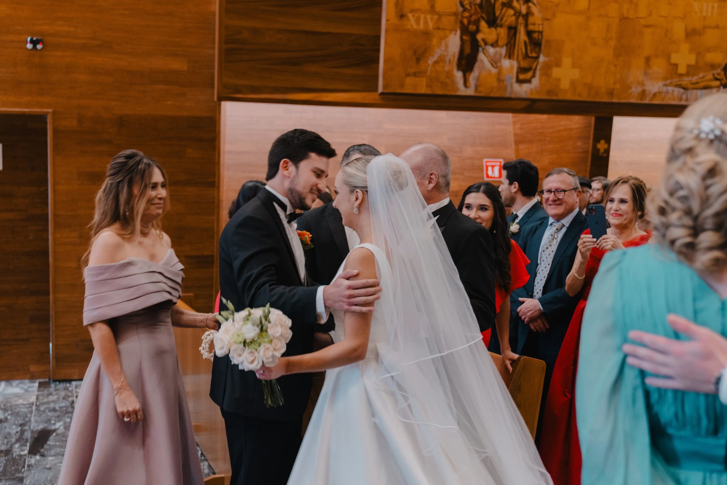 Una pareja de novios en su boda en una ceremonia religiosa, rodeados de familiares y amigos en una iglesia de madera, con rostros felices y un ambiente emotivo.