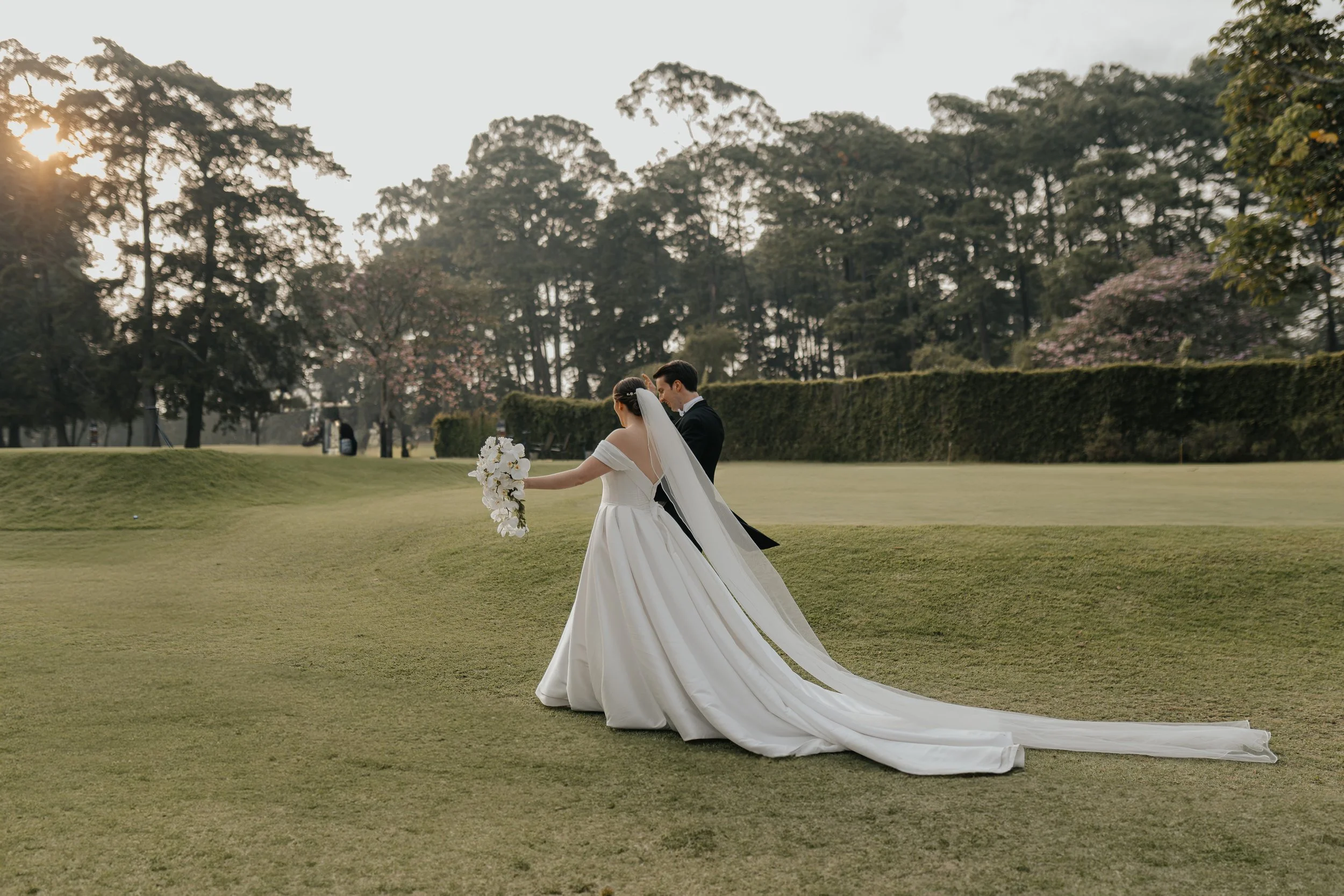 Pareja de novios en un campo de golf, la novia con vestido de boda y bouquet, el novio con frac, en la puesta del sol.