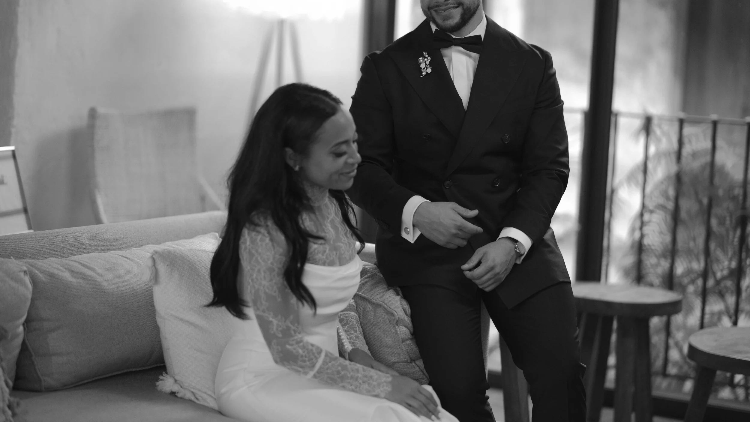 Hombre vestido en traje formal con pajarita ayudando a mujer en vestido de novia en un ambiente interior.