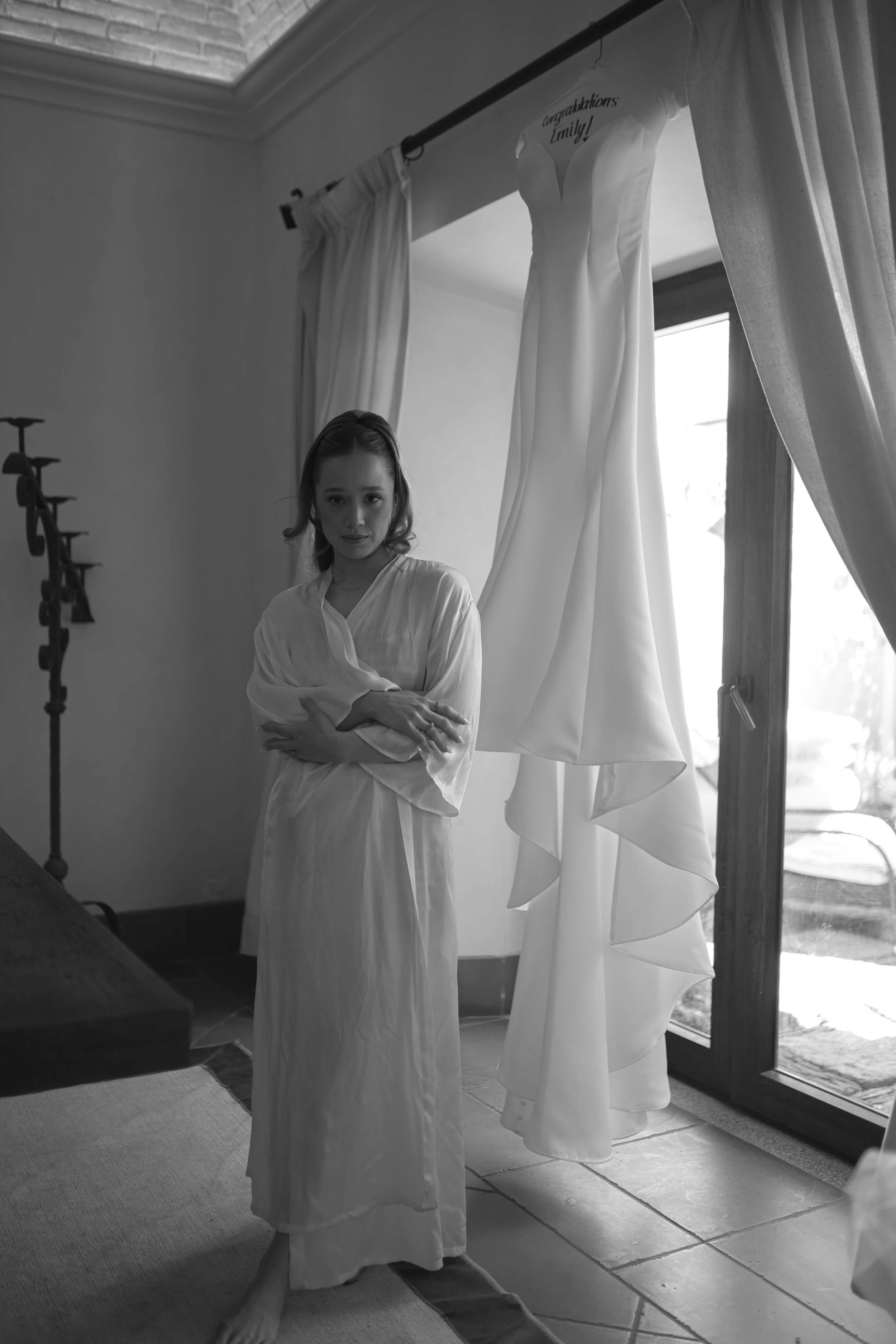 Mujer en vestido largo y con cabello rizado, de pie en una habitación junto a un vestido de novia colgado, con cortinas y una puerta de vidrio al fondo, en blanco y negro.