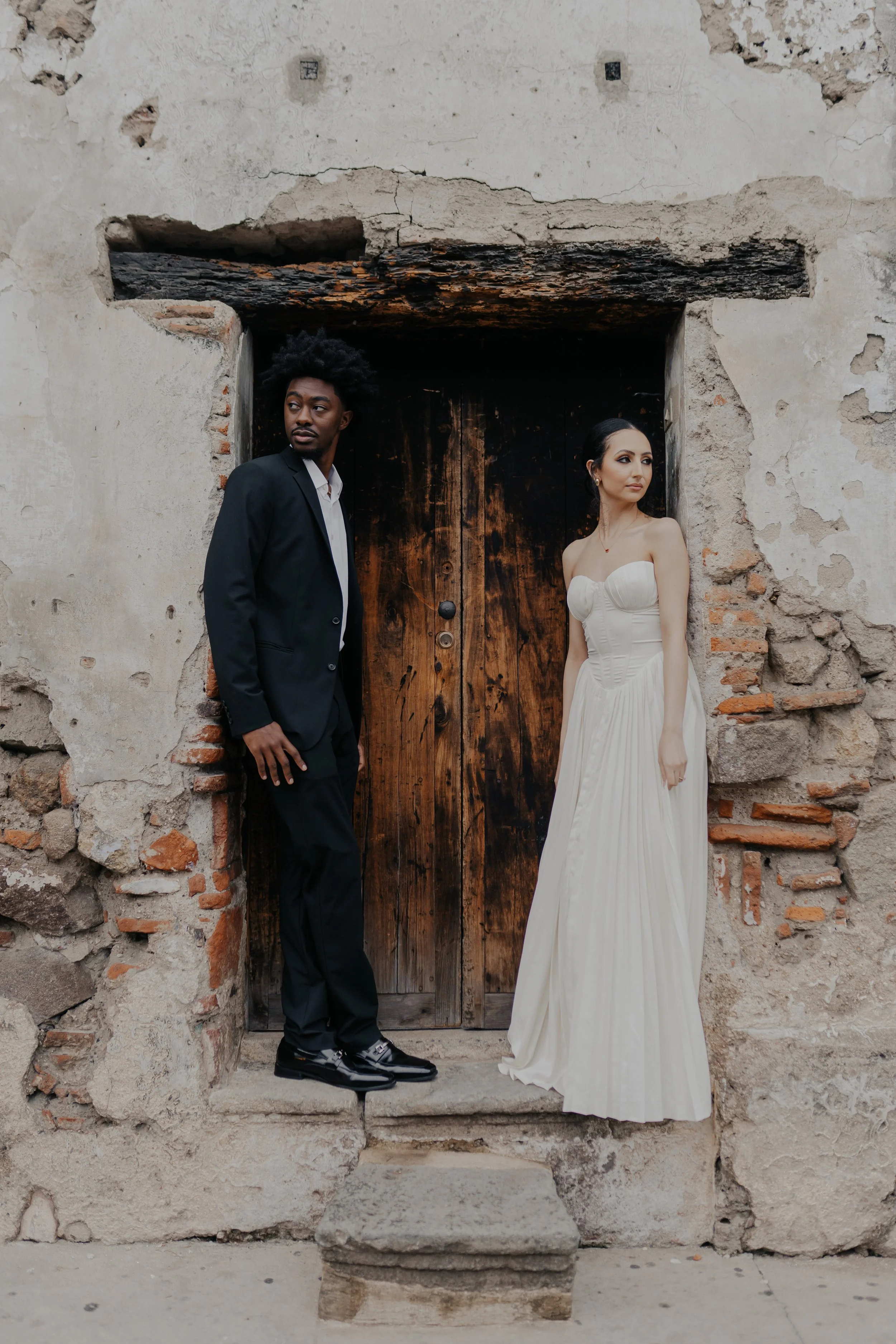 Una pareja vestida con ropa formal, un hombre en traje negro y una mujer en vestido largo de novia, posando frente a una puerta de madera en una pared de piedra antigua.