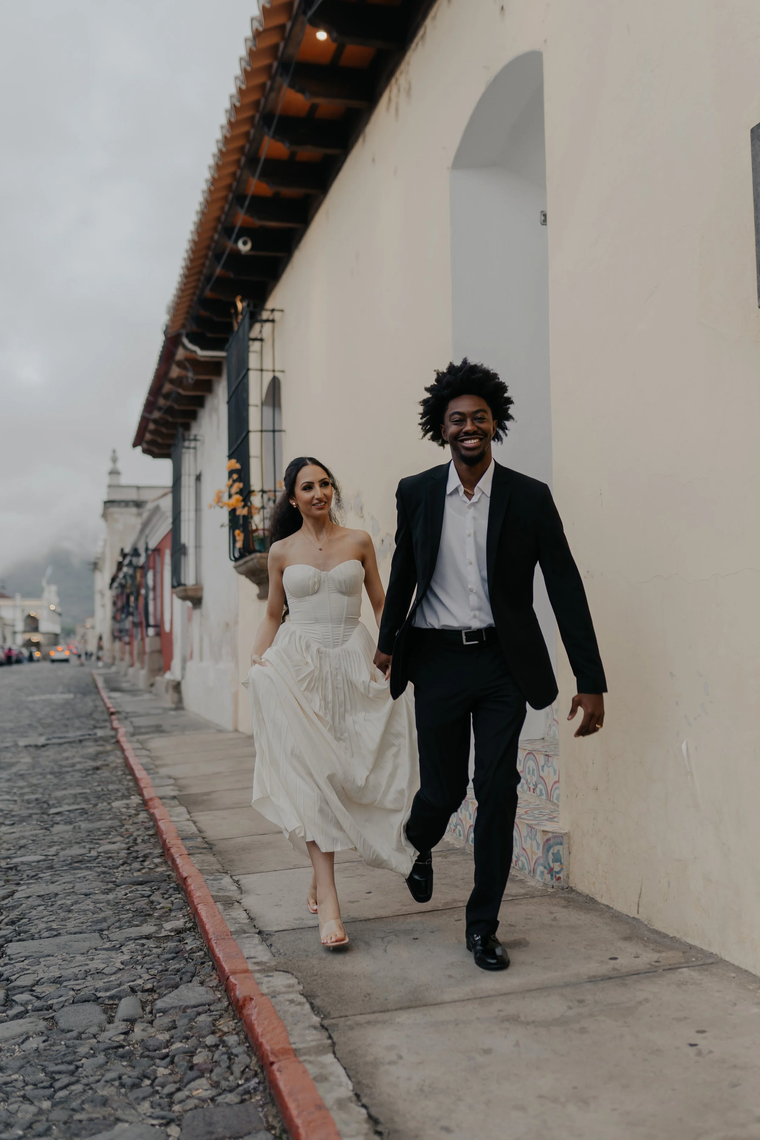 Pareja de novios caminando por una calle empedrada, ella en vestido de novia y él en traje, sonriendo y disfrutando del momento.