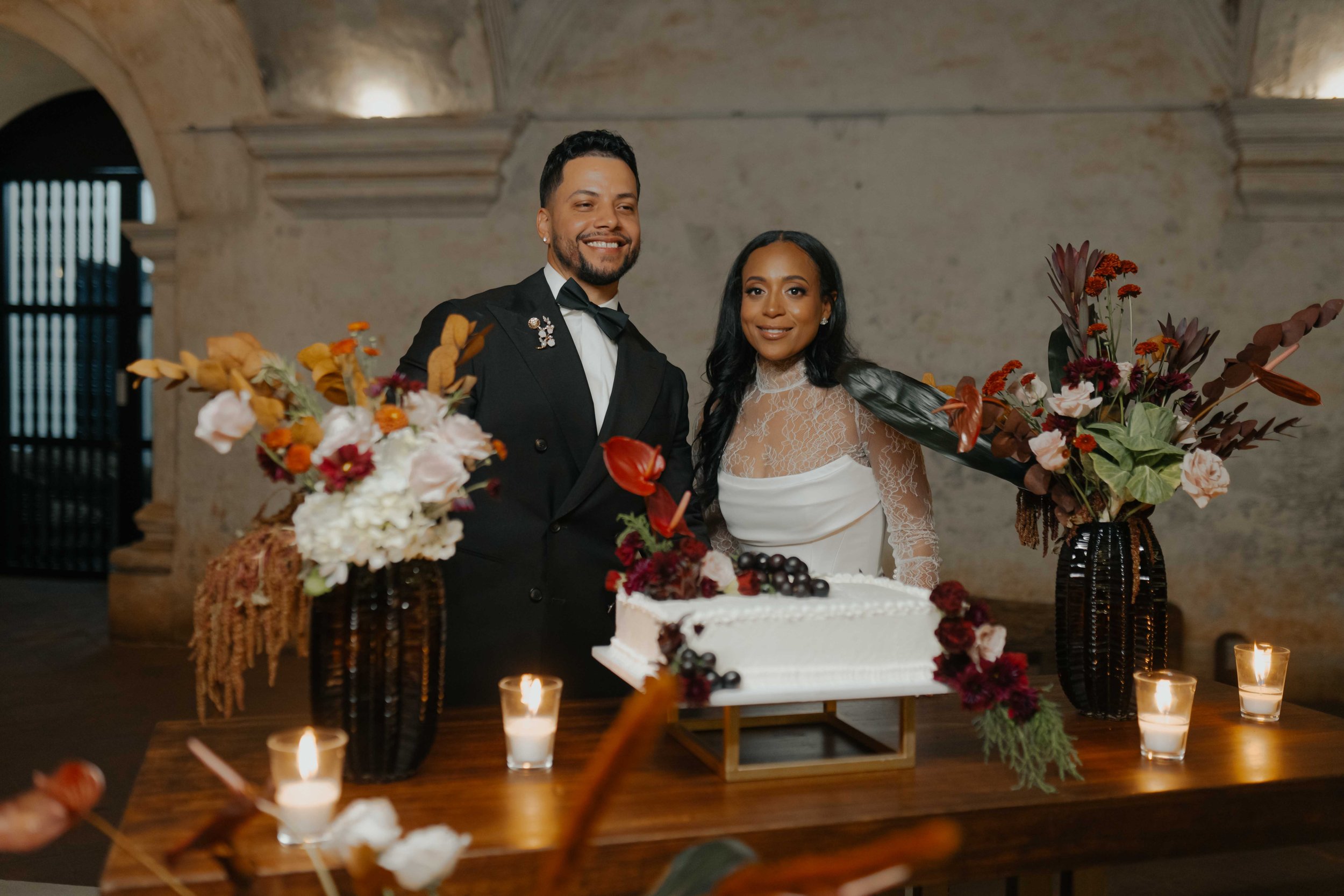 Pareja de novios en una boda, sonriendo detrás de una mesa con pastel decorado con frutas y velas, rodeados de arreglos florales variados en jarrones negros en un lugar con paredes de piedra.