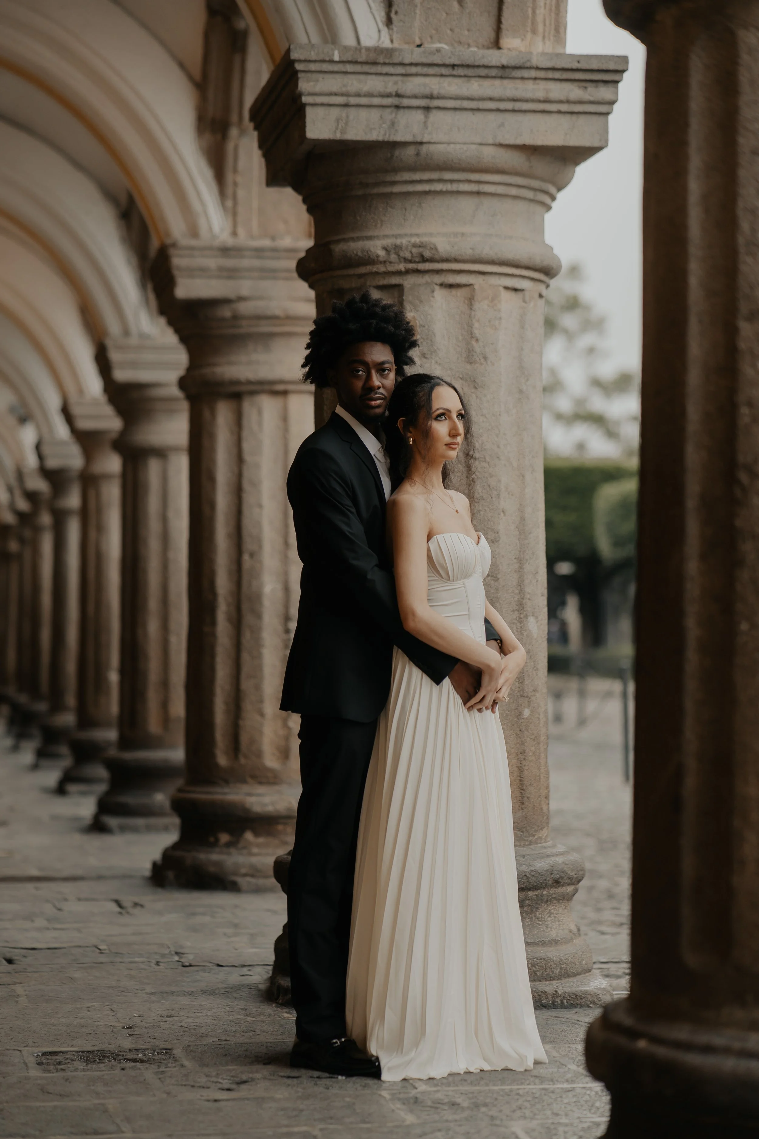 Pareja en vestido de boda y traje formal, posando entre columnas de piedra en un lugar histórico o de arquitectura clásica.