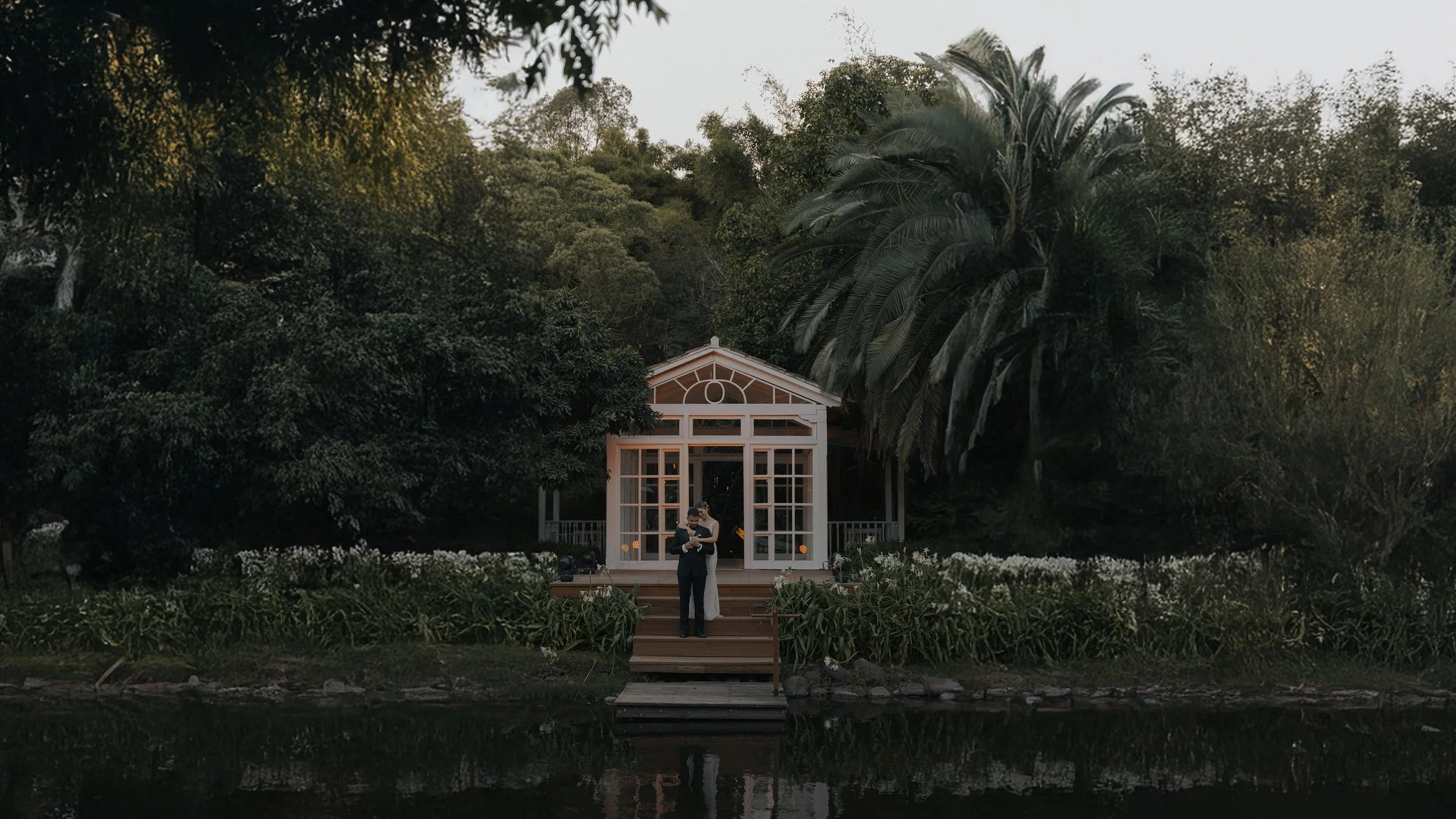 Pareja de novios en ceremonia en un pabellón en un entorno natural con árboles y agua.