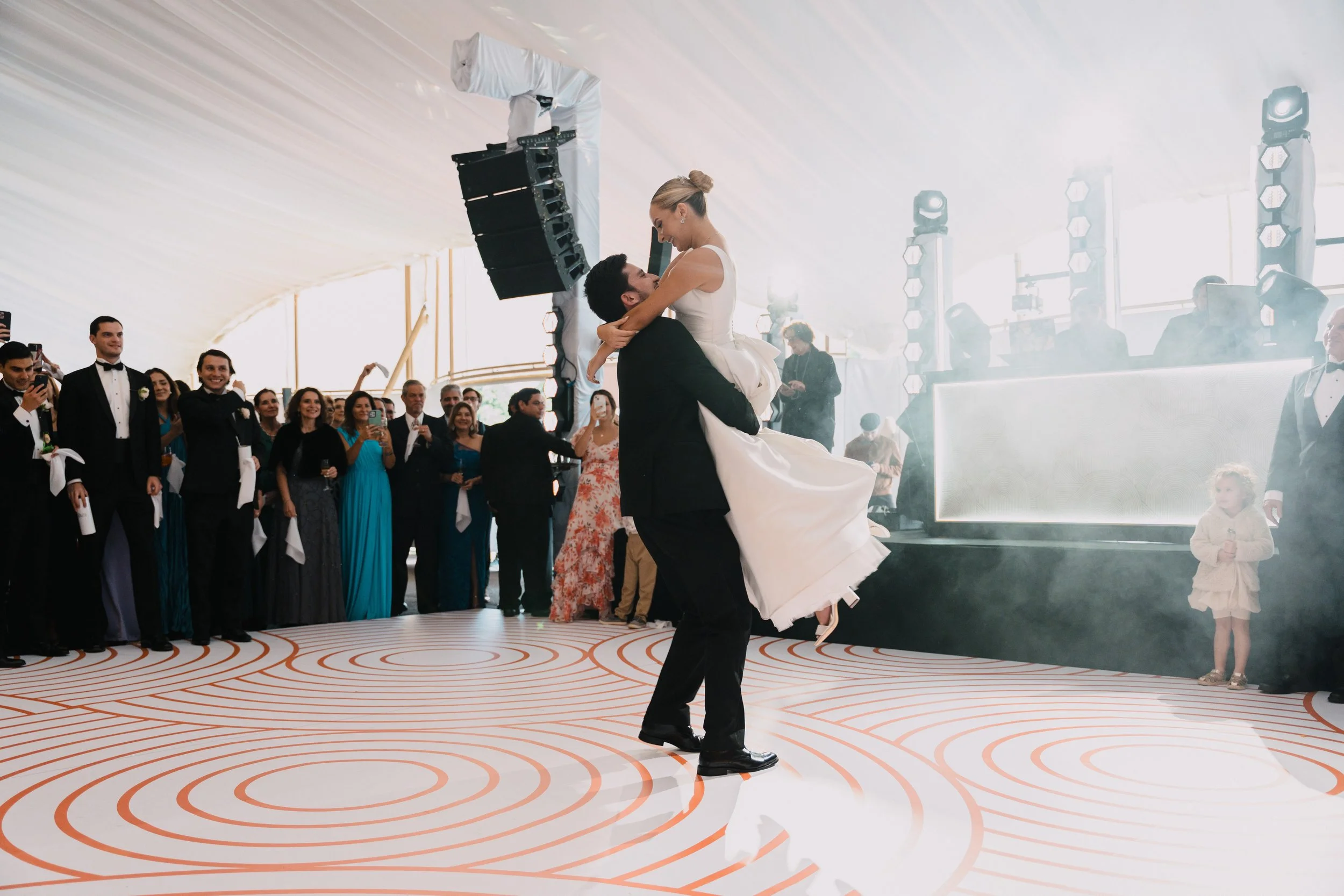 Pareja bailando en una fiesta de bodas, con invitados observando y una pista de baile decorada.