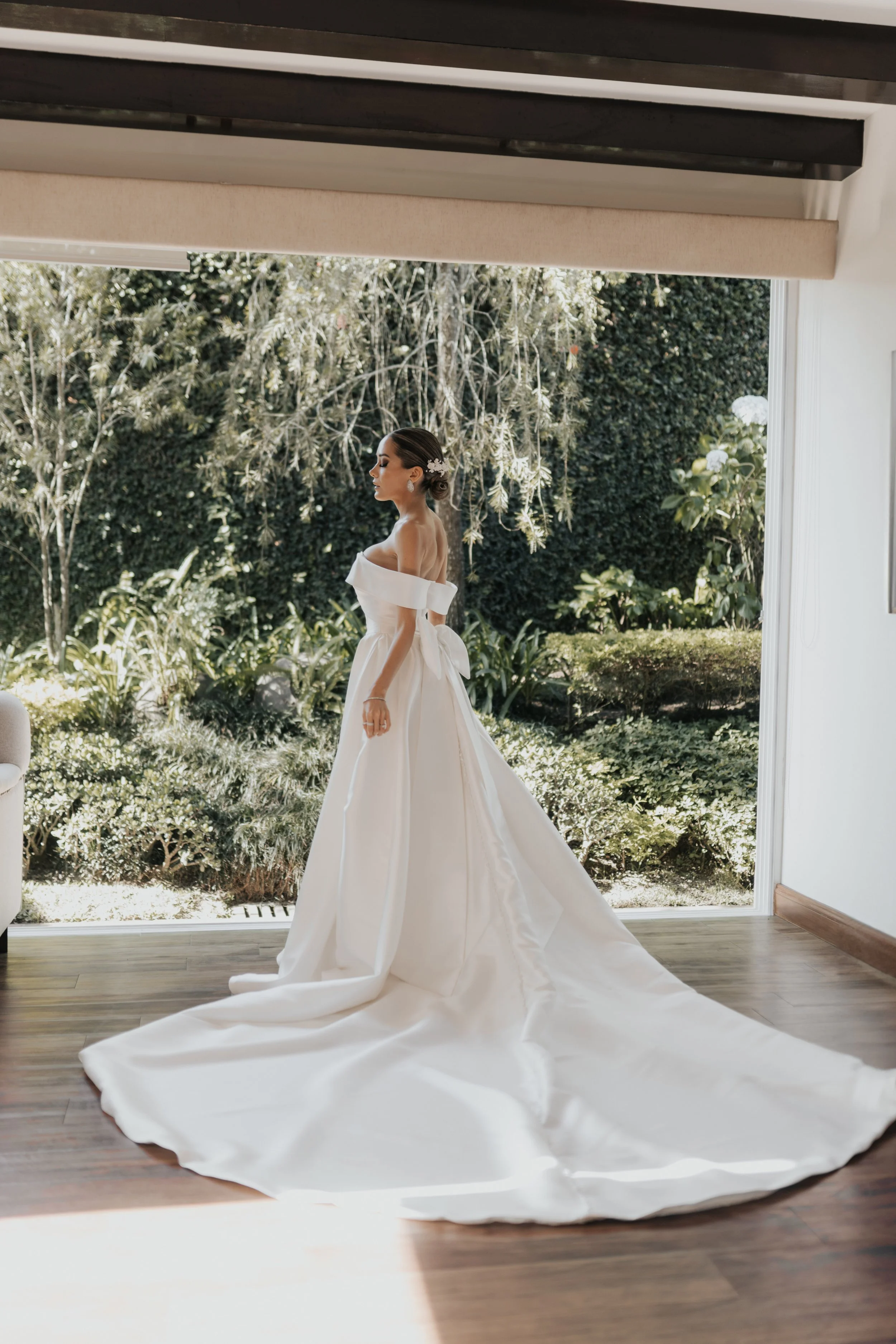 Una mujer con vestido de novia blanco de encaje y cola larga en una habitación con ventana que muestra un jardín con árboles y plantas.