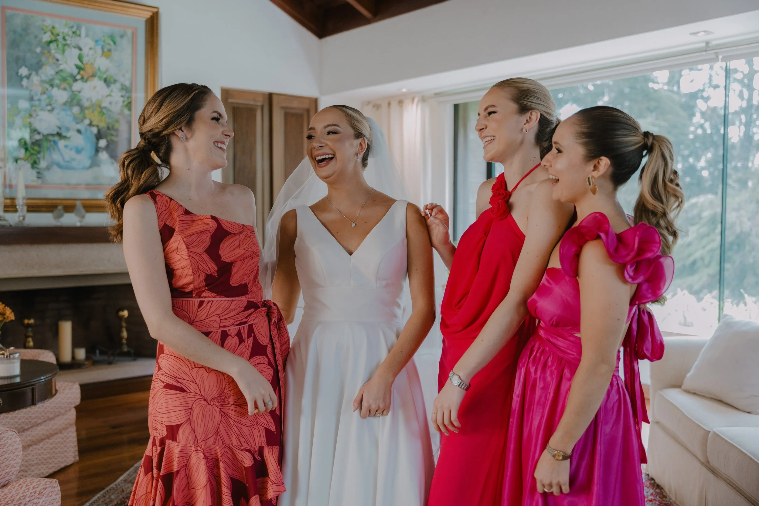 Cinco mujeres sonriendo y riendo en una sala con ventanas y decoración moderna. Una mujer de novia con vestido blanco y velo, acompañada por cuatro mujeres con vestidos de diferentes colores y estilos, en un momento alegre y emotivo.