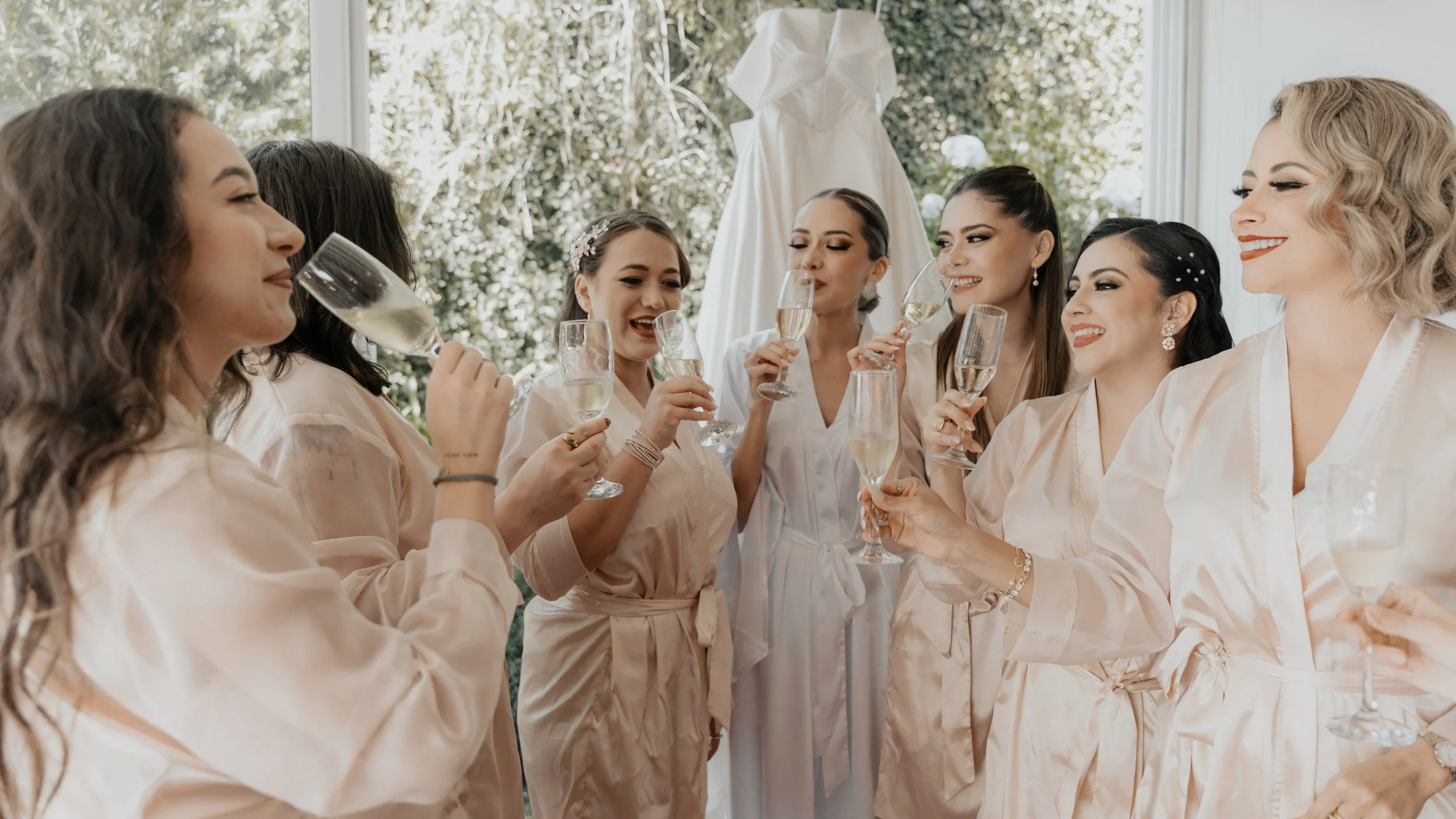 Grupo de mujeres en bata de seda brindando con copas de champán en una celebración, con vestido de novia colgado en el fondo