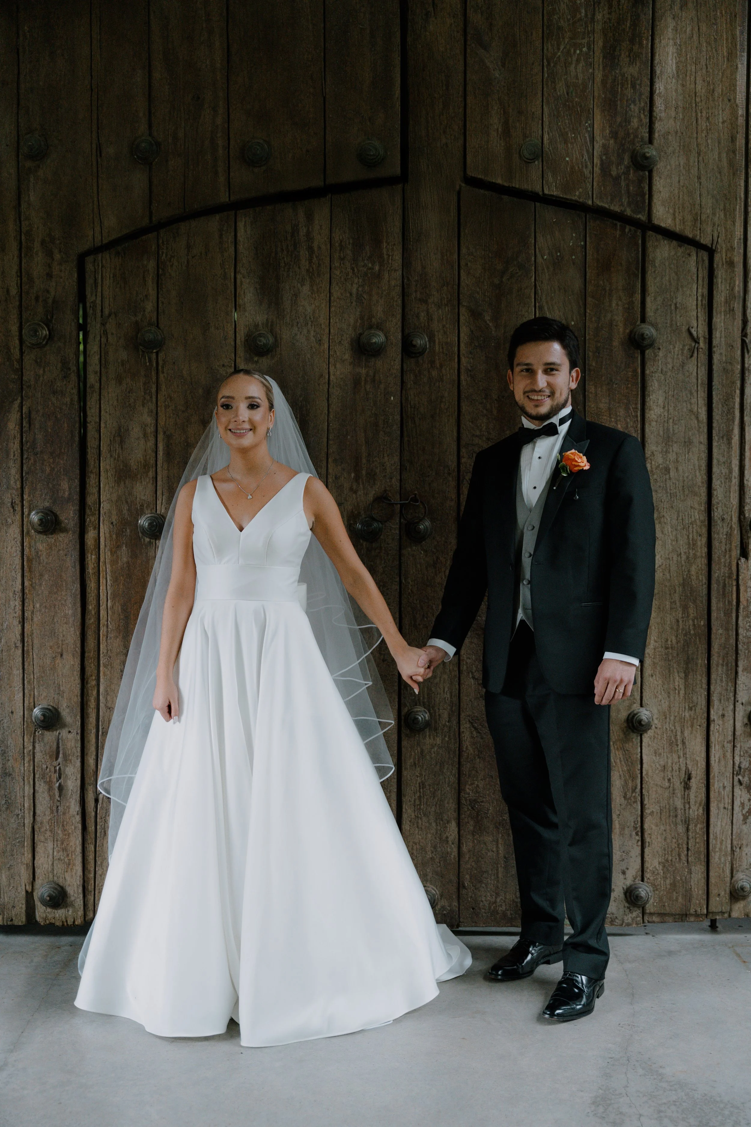 Una pareja de recién casados sosteniendo las manos frente a una puerta de madera grande, ella lleva vestido de novia blanco y velo, él lleva traje negro con pajarita y boutonnière naranja.