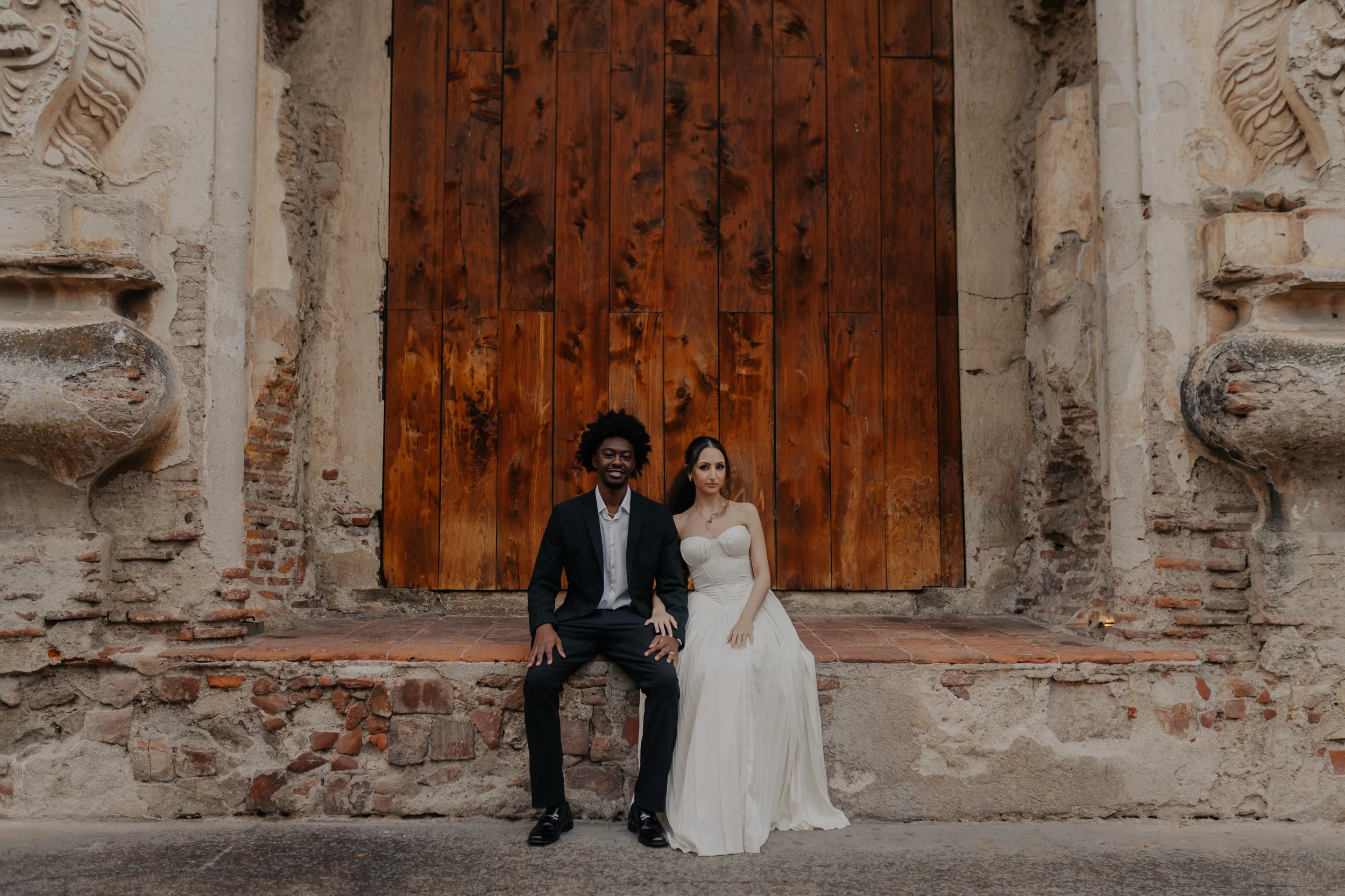 Una pareja vestida con ropa formal sentada en un escalón frente a una puerta de madera grande y antigua, rodeada de muros de piedra y ladrillo.