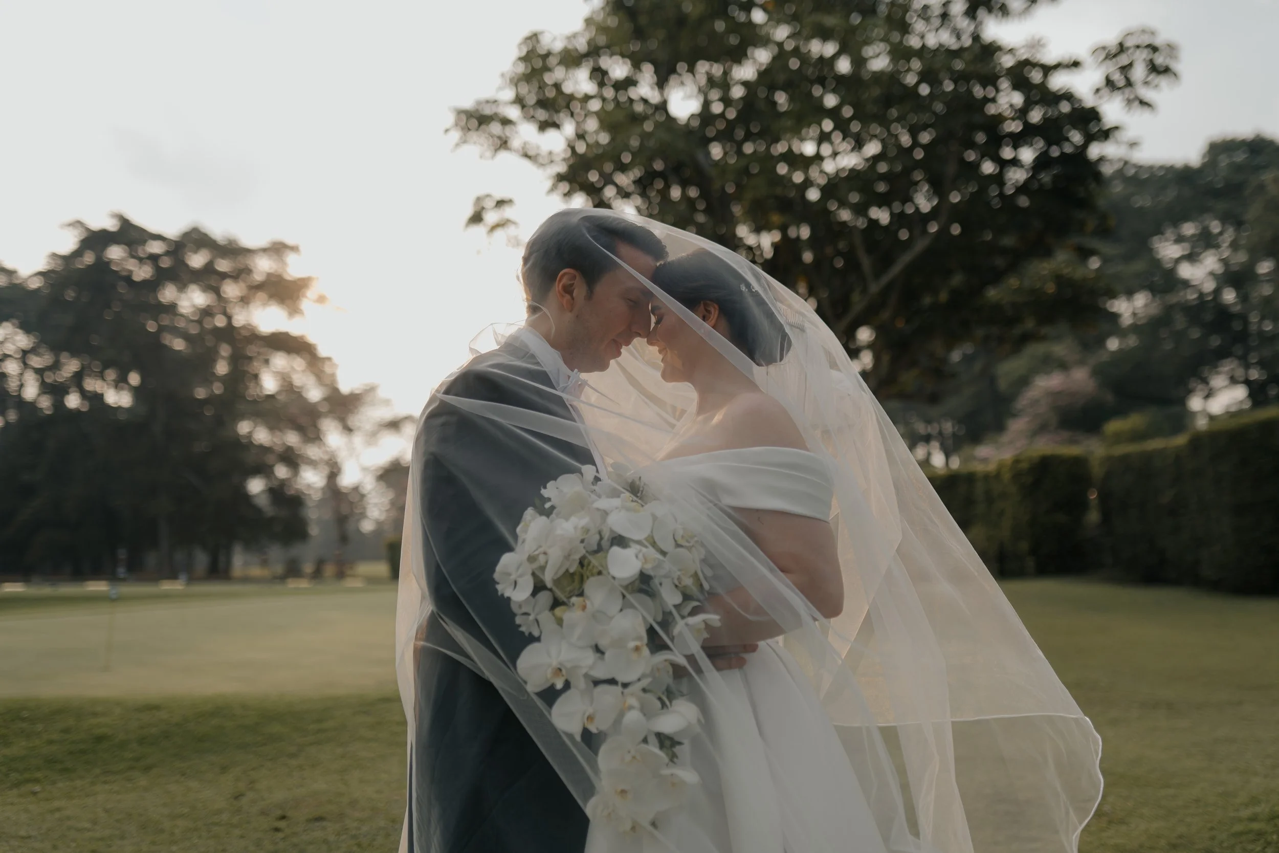 Pareja de recién casados en un campo, la novia con velo y vestido blanco, sosteniendo un ramo de flores blancas, con árboles al fondo y puesta de sol.
