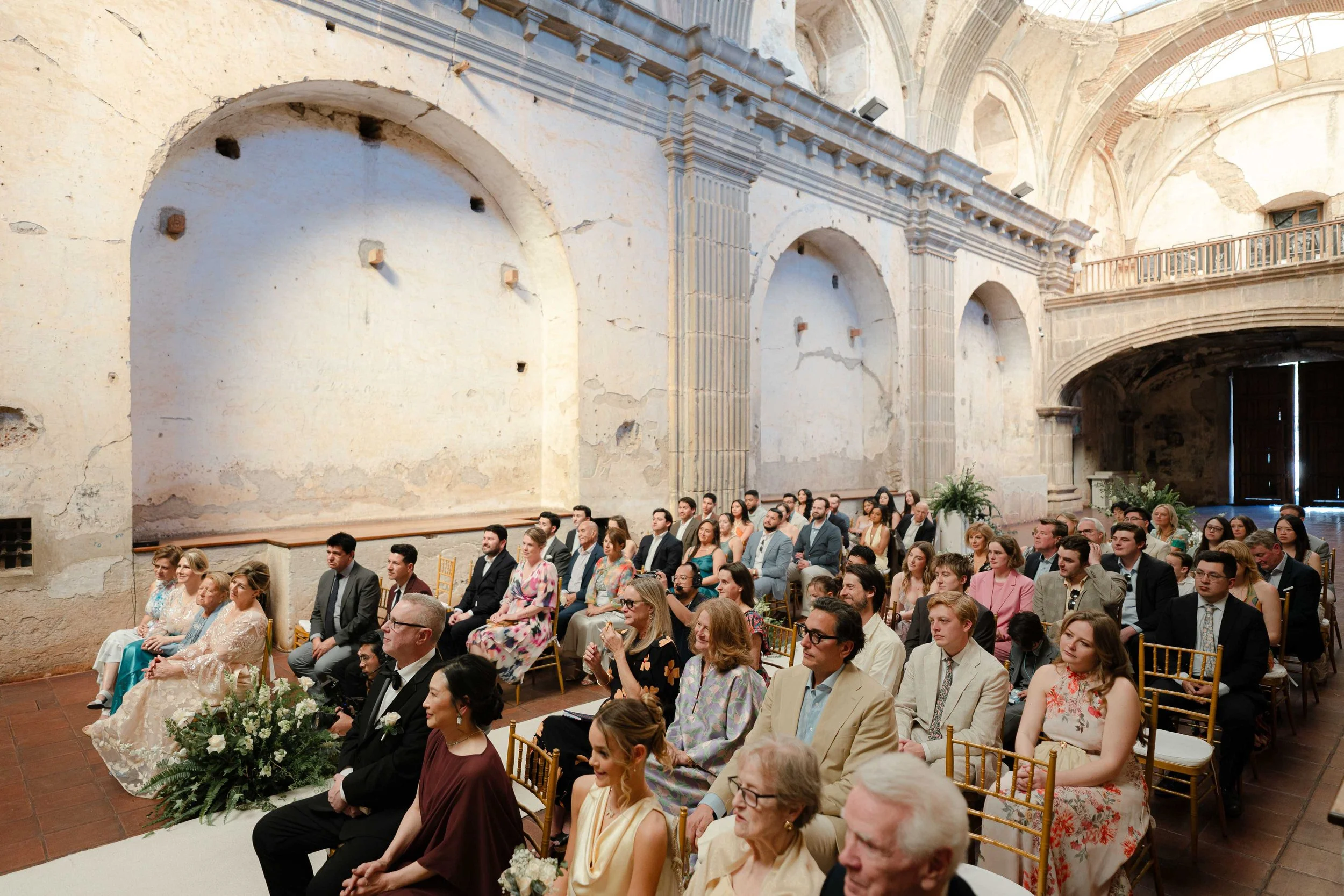 Una ceremonia de boda en un lugar histórico con paredes de piedra y arcos, con muchas personas sentadas mirando hacia adelante.