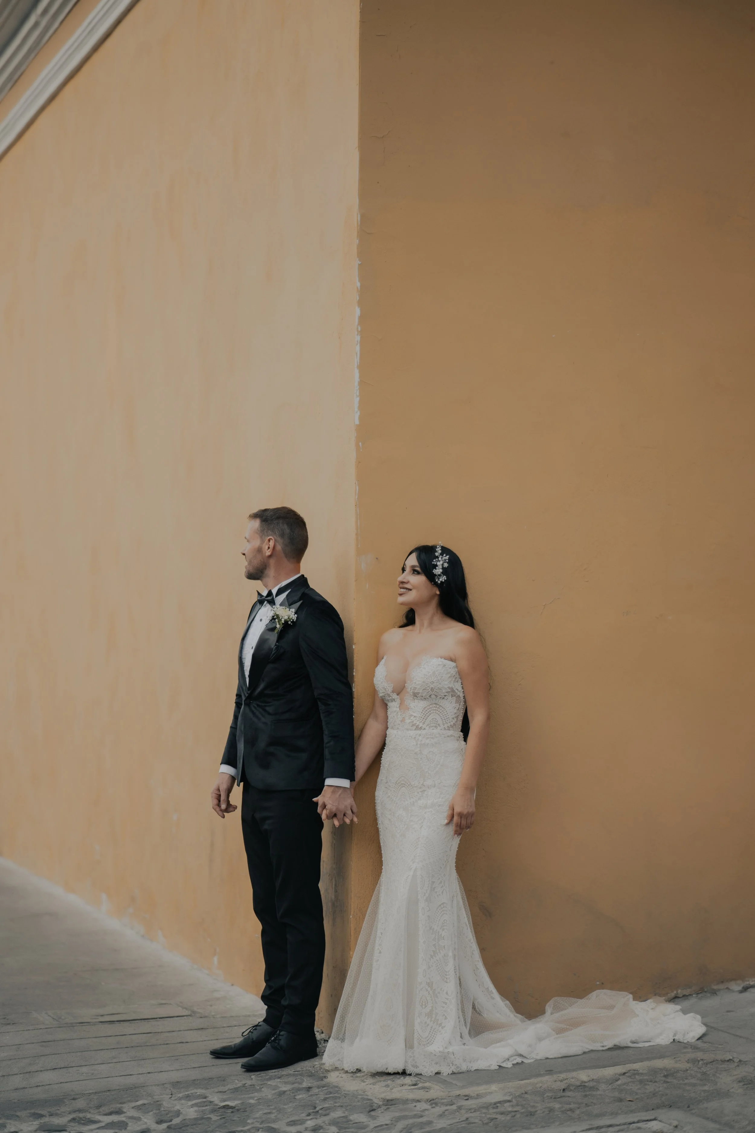 Una pareja de novios de espaldas, con las manos entrelazadas, apoyados en una pared anaranjada, en una boda.