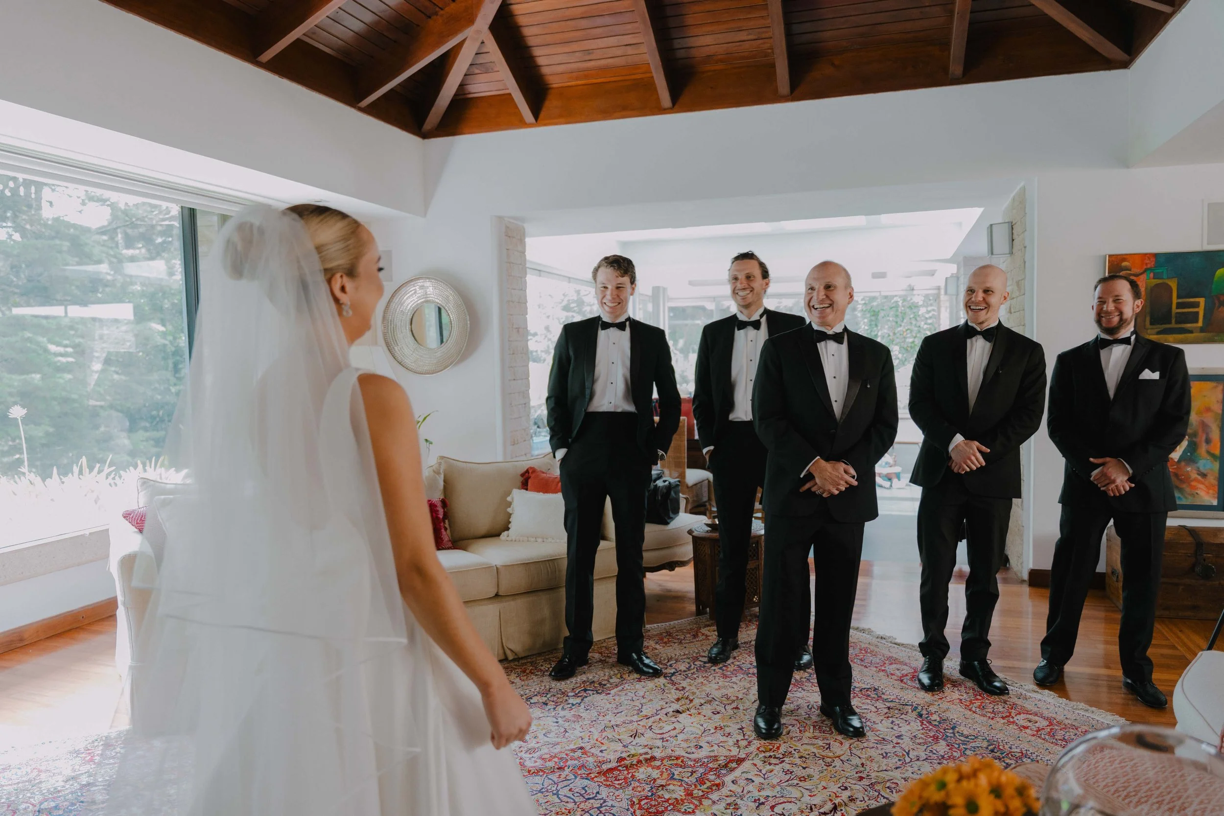 Mujer vestida de novia frente a cinco hombres de traje en una sala luminosa y moderna con decoración artística.