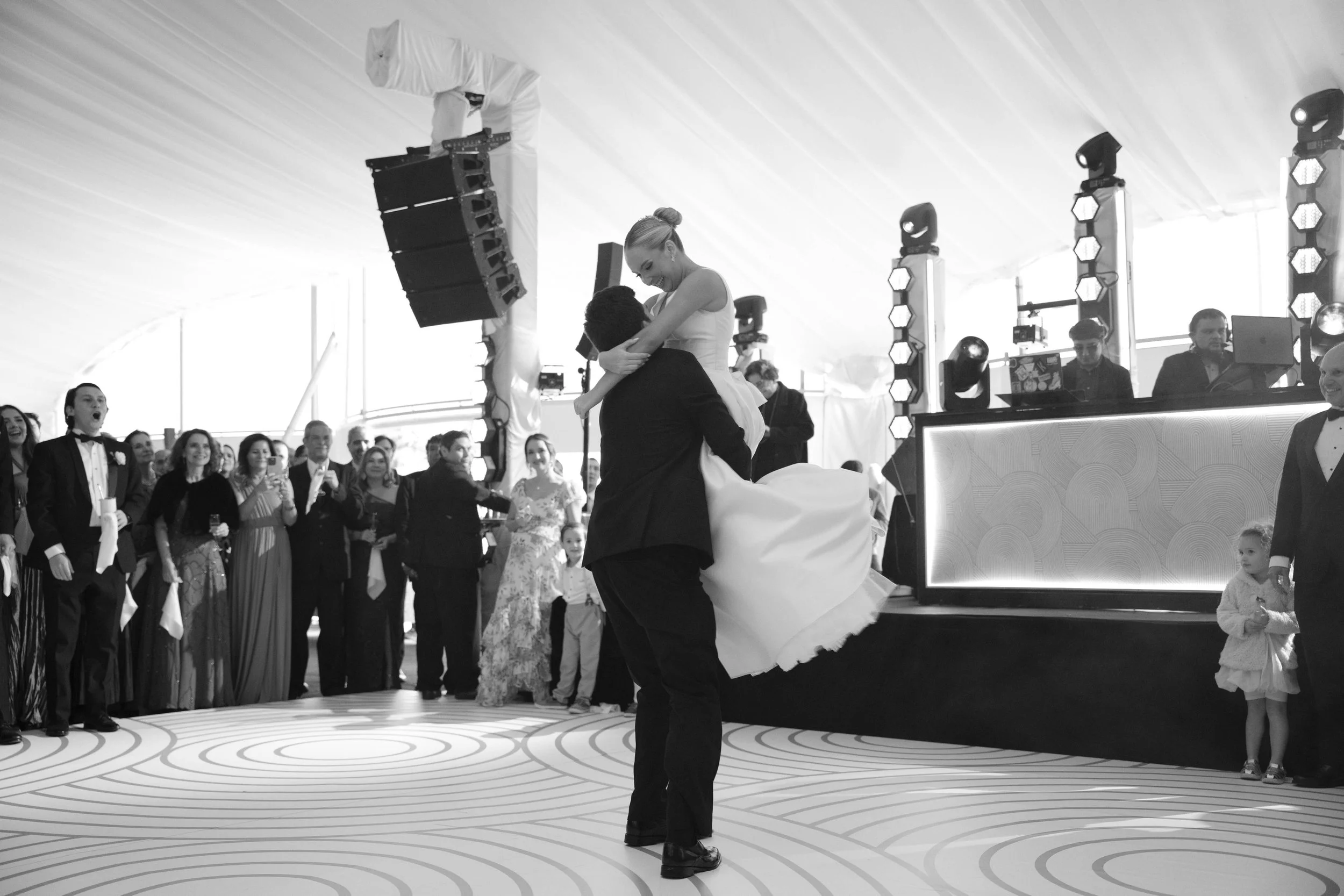 Pareja bailando en una celebración de boda, rodeada de invitados y un DJ o banda en un escenario.
