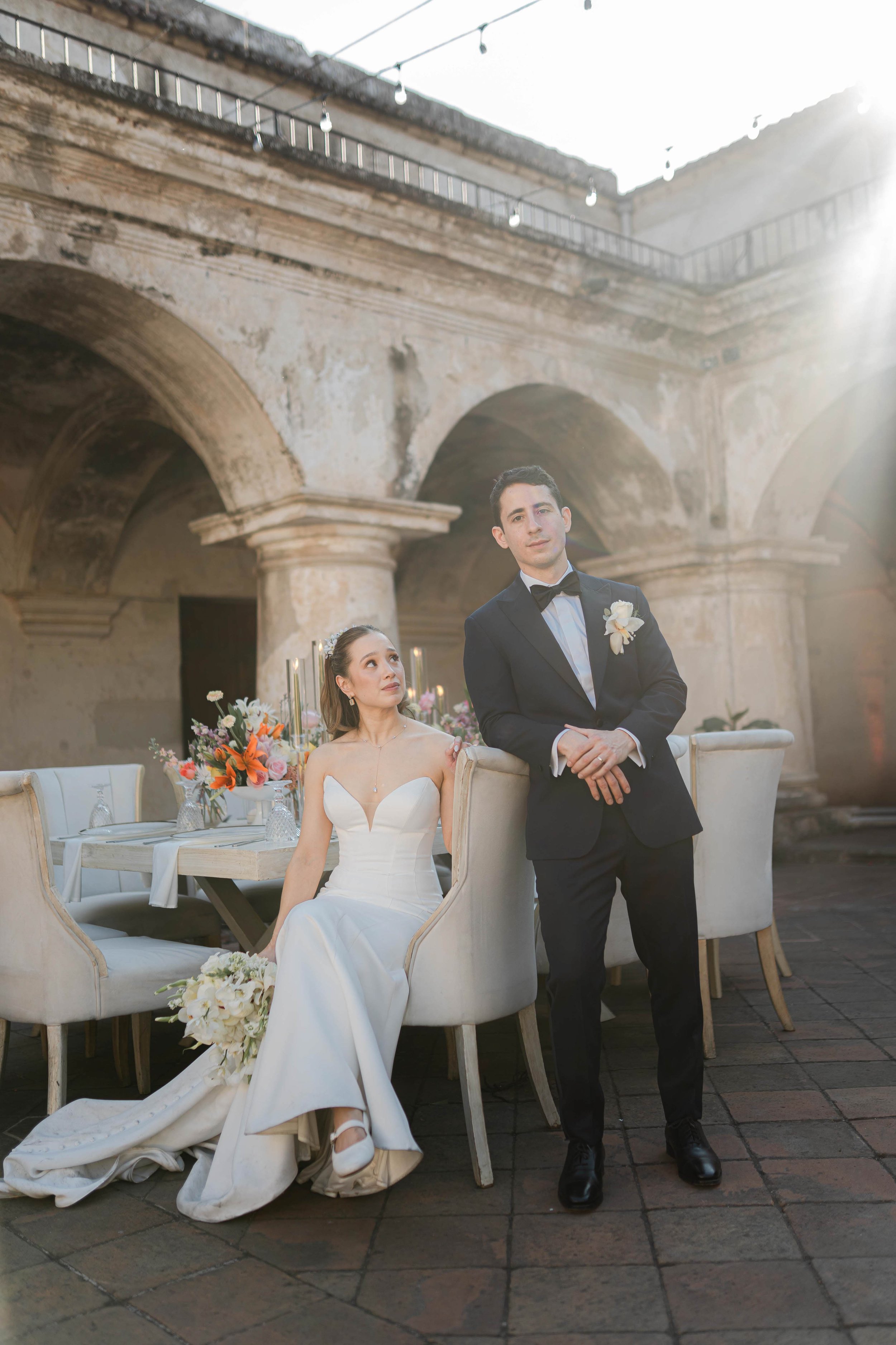 Pareja de recién casados en una boda en un patio con arquitectura antigua, decoración con flores y velas, luz solar brillante.