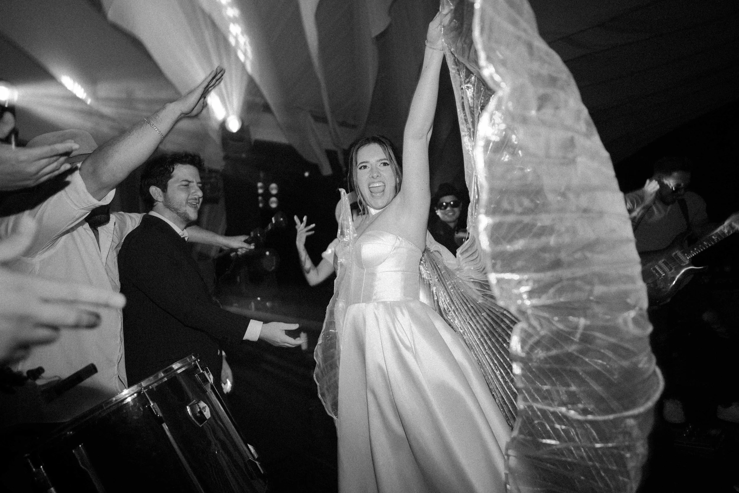 Mujer vestida con vestido de novia, bailando con entusiasmo en una fiesta, rodeada de amigos que también bailan y disfrutan del momento, en un evento festivo con decoración y música en vivo.
