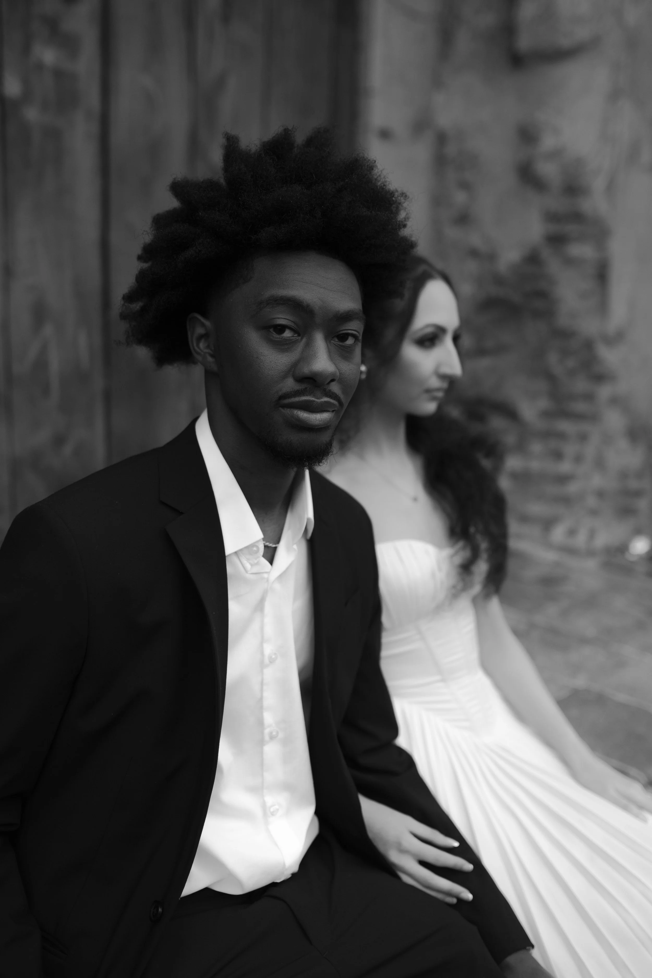 Un hombre y una mujer en un entorno de piedra, con el hombre en primer plano y la mujer al fondo, ambos vestidos formalmente.