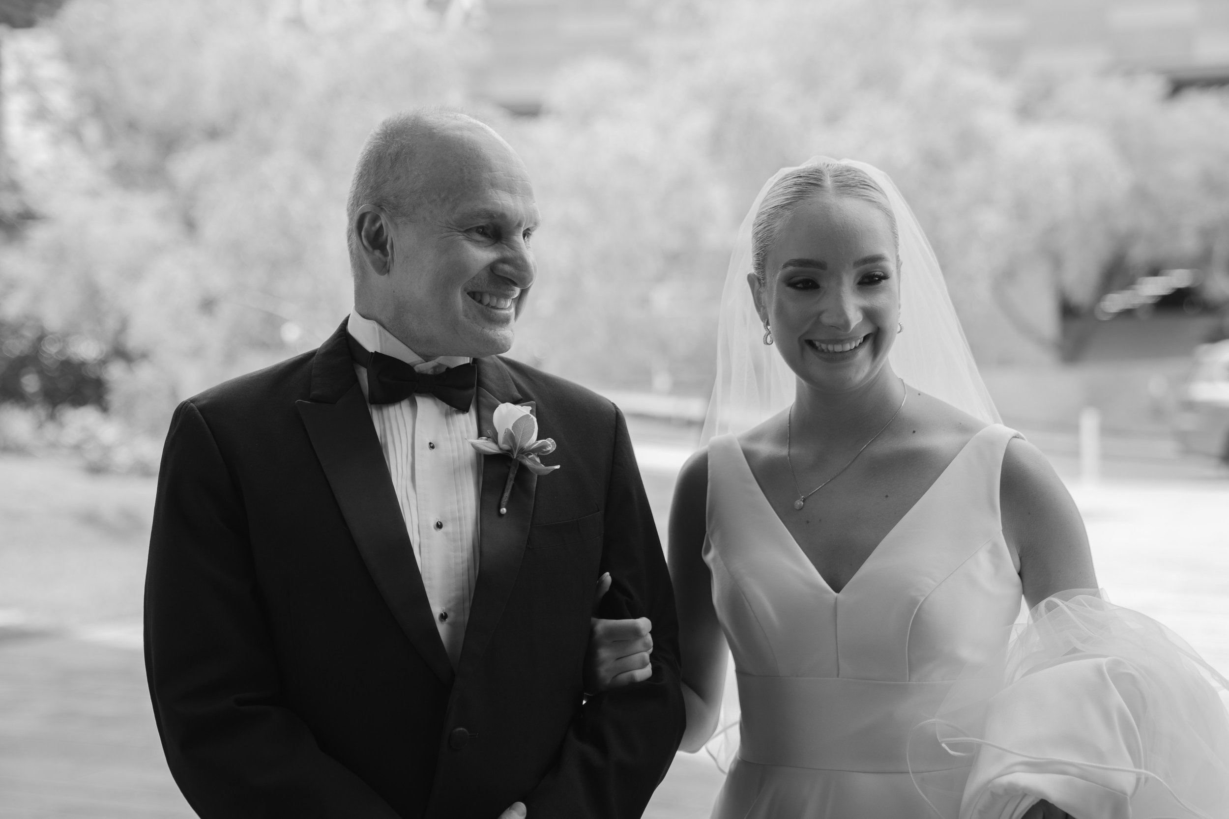 Una escena de bodas en blanco y negro con una mujer vestida de novia y un hombre vestido con traje formal. La novia lleva vestido blanco con fondo en V, velo y accesorios de joyería, y la pareja se ve feliz.