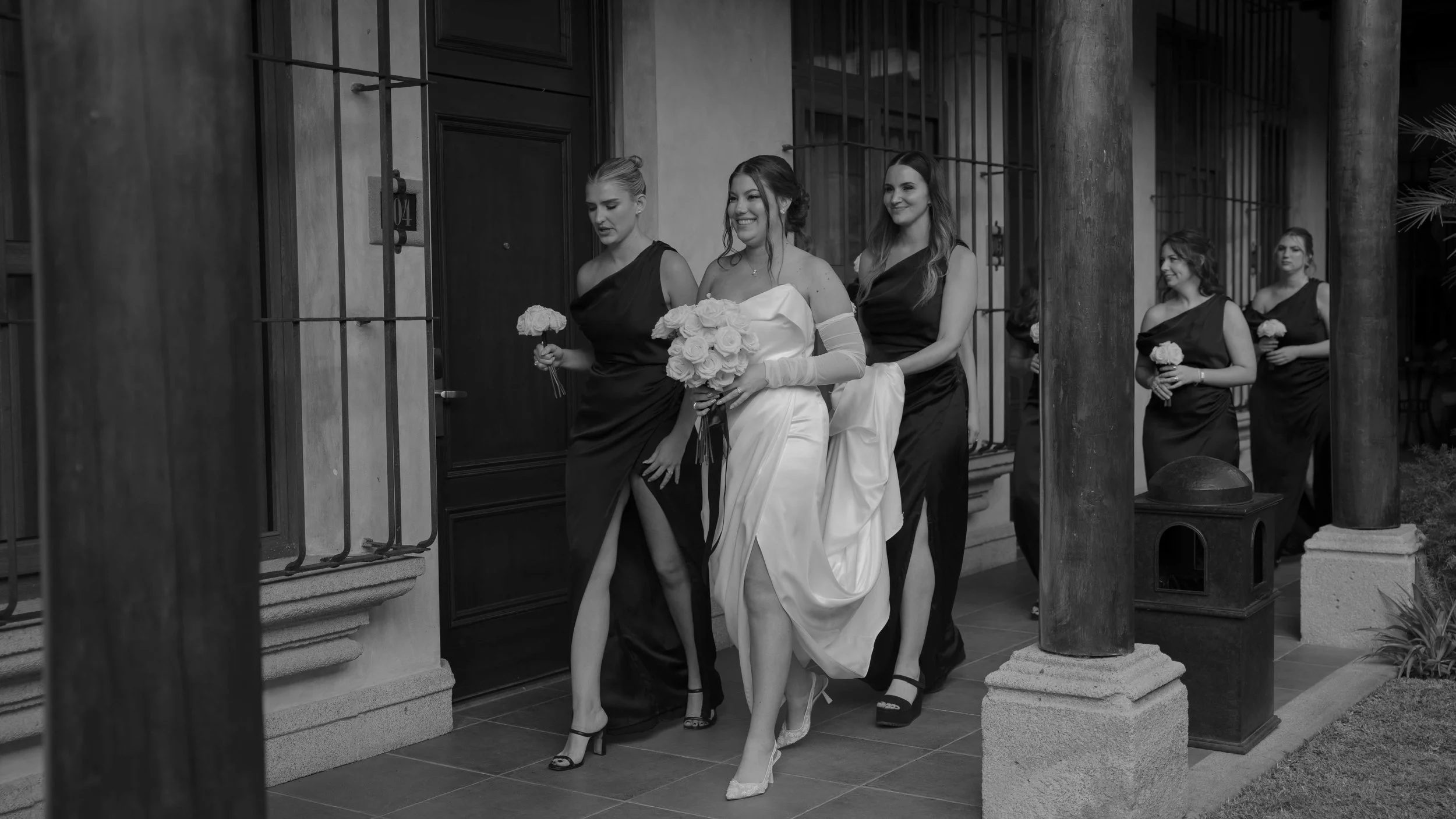 Mujer vestida de novia con ramo de rosas y cinco damas de honor con ramos, caminando por un porche con columnas de piedra y rejas negras.