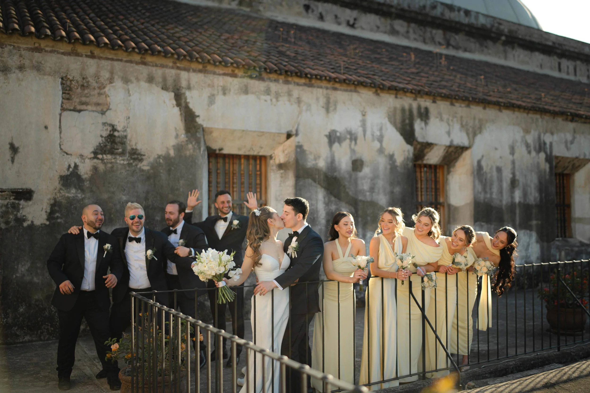 Grupo de personas en una celebración de boda al aire libre, con la novia y el novio besándose en el centro y los invitados sonriendo y haciendo gestos de alegría, en un fondo de paredes antiguas y un techo de tejas.