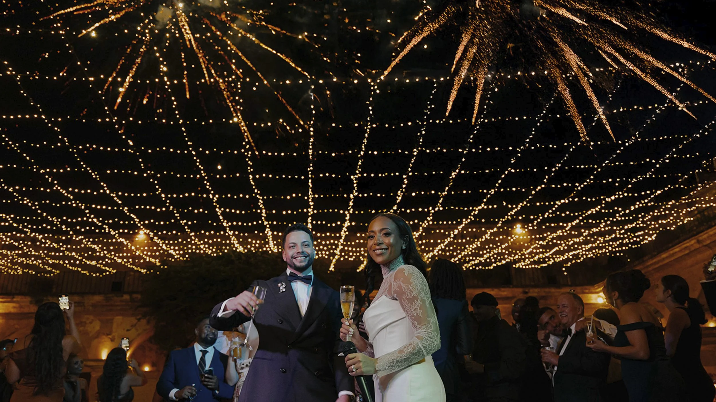 Pareja de novios en una boda con decoración de luces doradas en el techo, rodeados de invitados que celebran con copas en un ambiente festivo y elegante.