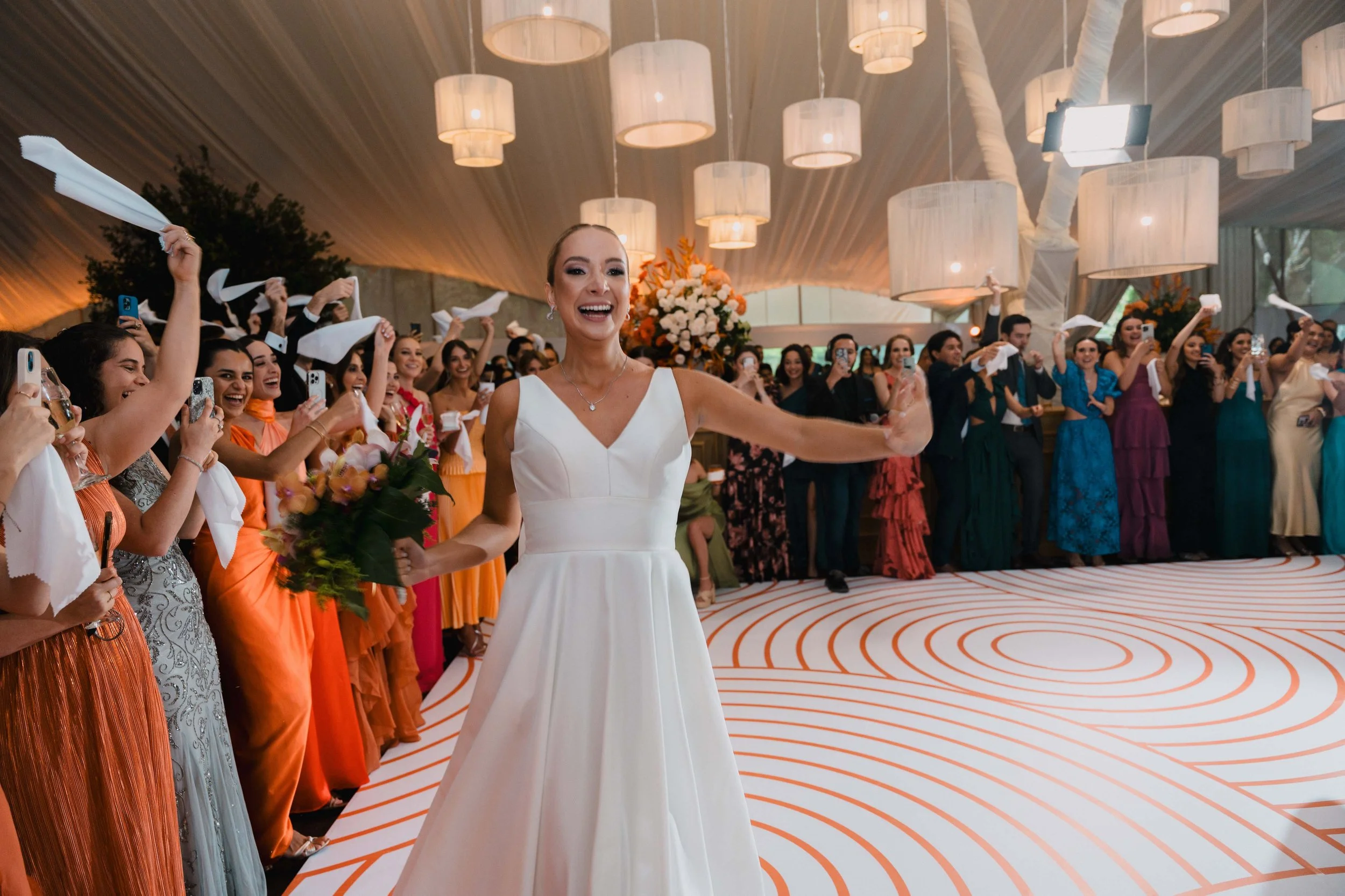 Mujer de vestido blanco en una boda, rodeada de invitados felices, algunos lanzando servilletas y otros tomando fotos, en un salón decorado con luces y flores.