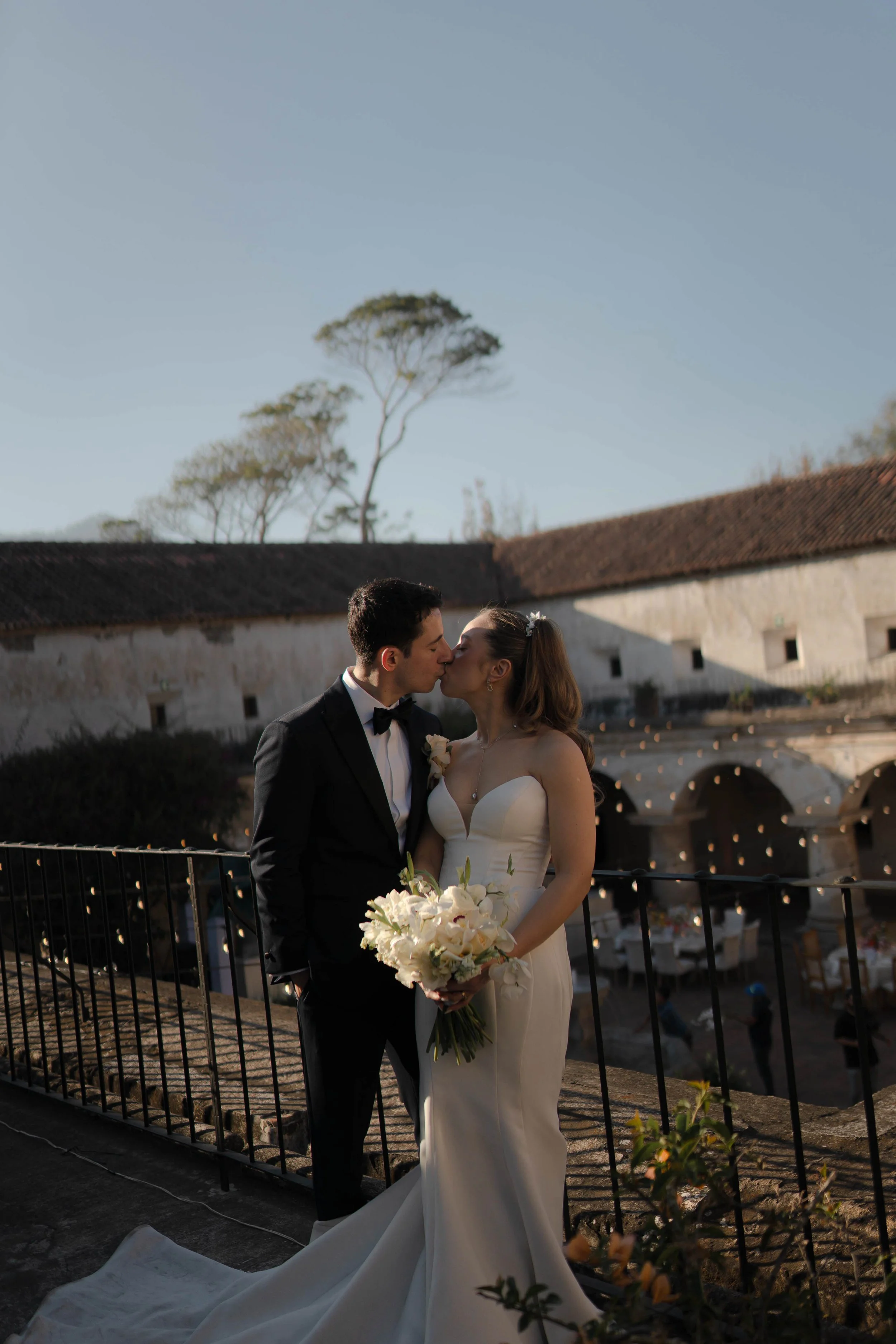 Una pareja de novios besándose en una boda al aire libre, la novia lleva un vestido blanco y sostiene un ramo de flores blancas, el novio viste un traje negro con pajarita, en el fondo se ve un edificio antiguo y mesas para la recepción nupcial.