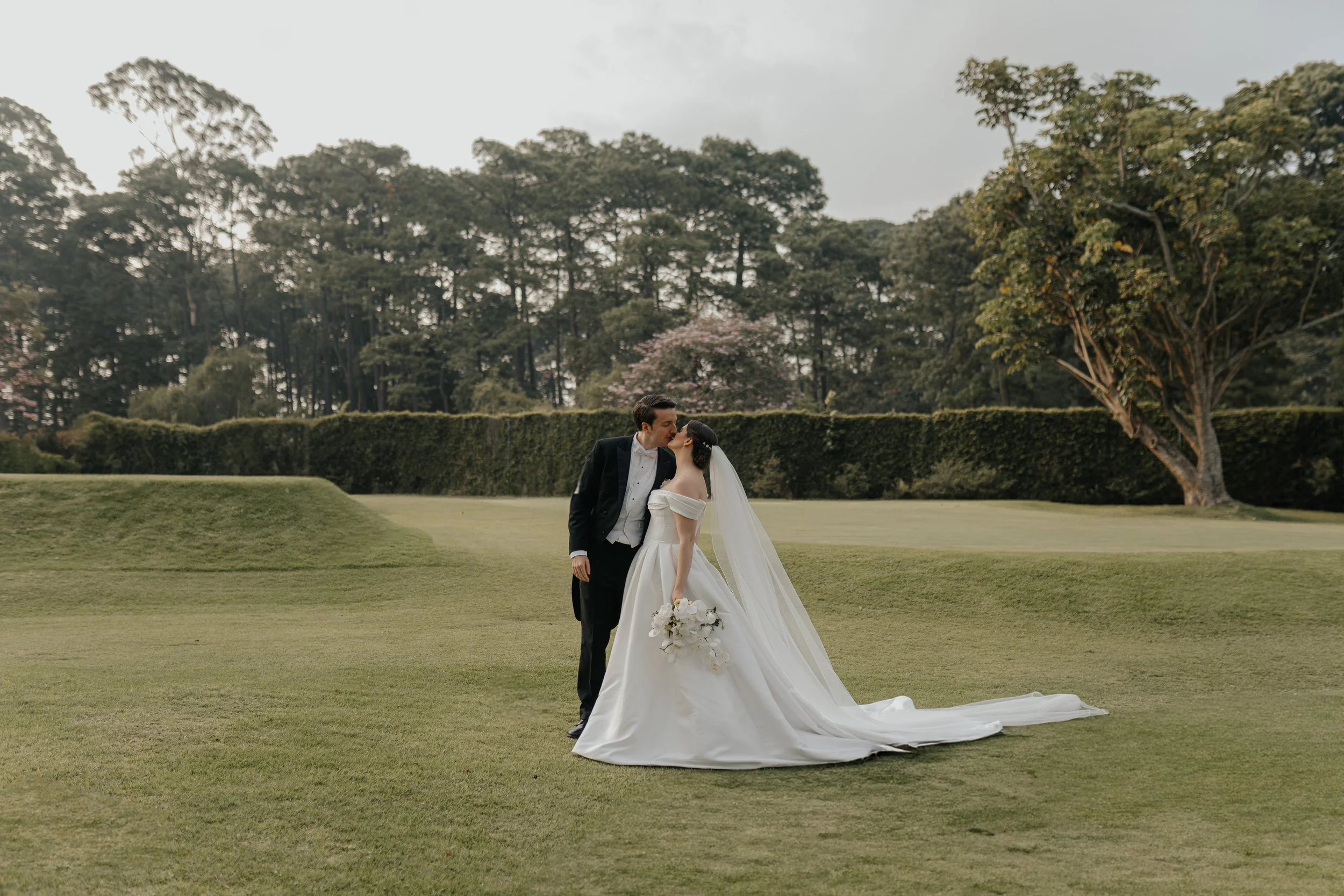 Pareja de novios en boda, la novia con vestido largo y velo, el novio con traje negro, en un campo con árboles y césped.