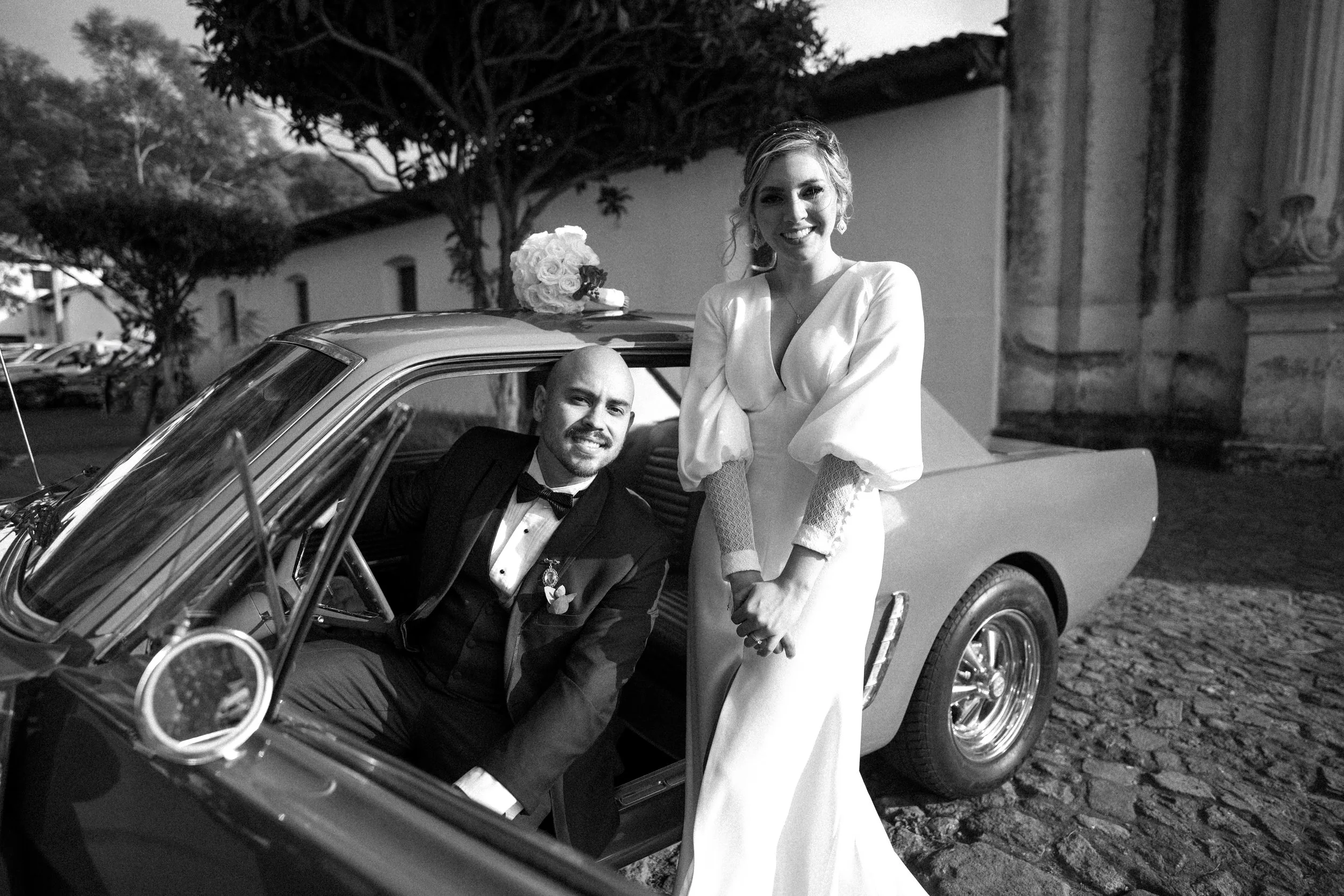 Fotografía en blanco y negro de una pareja en un coche clásico, con el hombre sentado en el coche y la mujer de pie junto a él, ambos vestidos con ropa formal, en un ambiente exterior con árboles y edificio histórico en el fondo, durante una celebrac